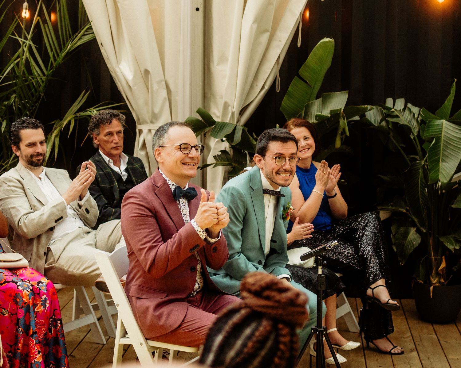 Guests clapping and smiling at an elegant indoor wedding ceremony surrounded by tropical plants.