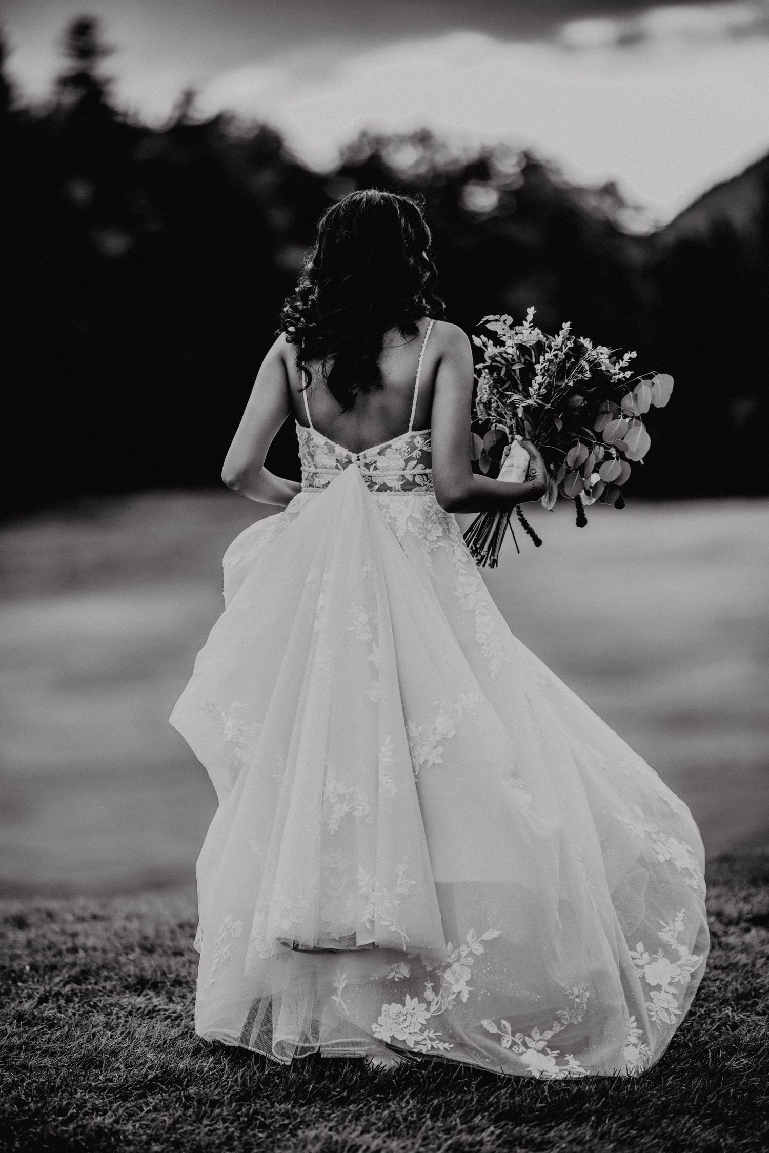 Bride in lace wedding gown holding floral bouquet, back view, black and white outdoor photo.