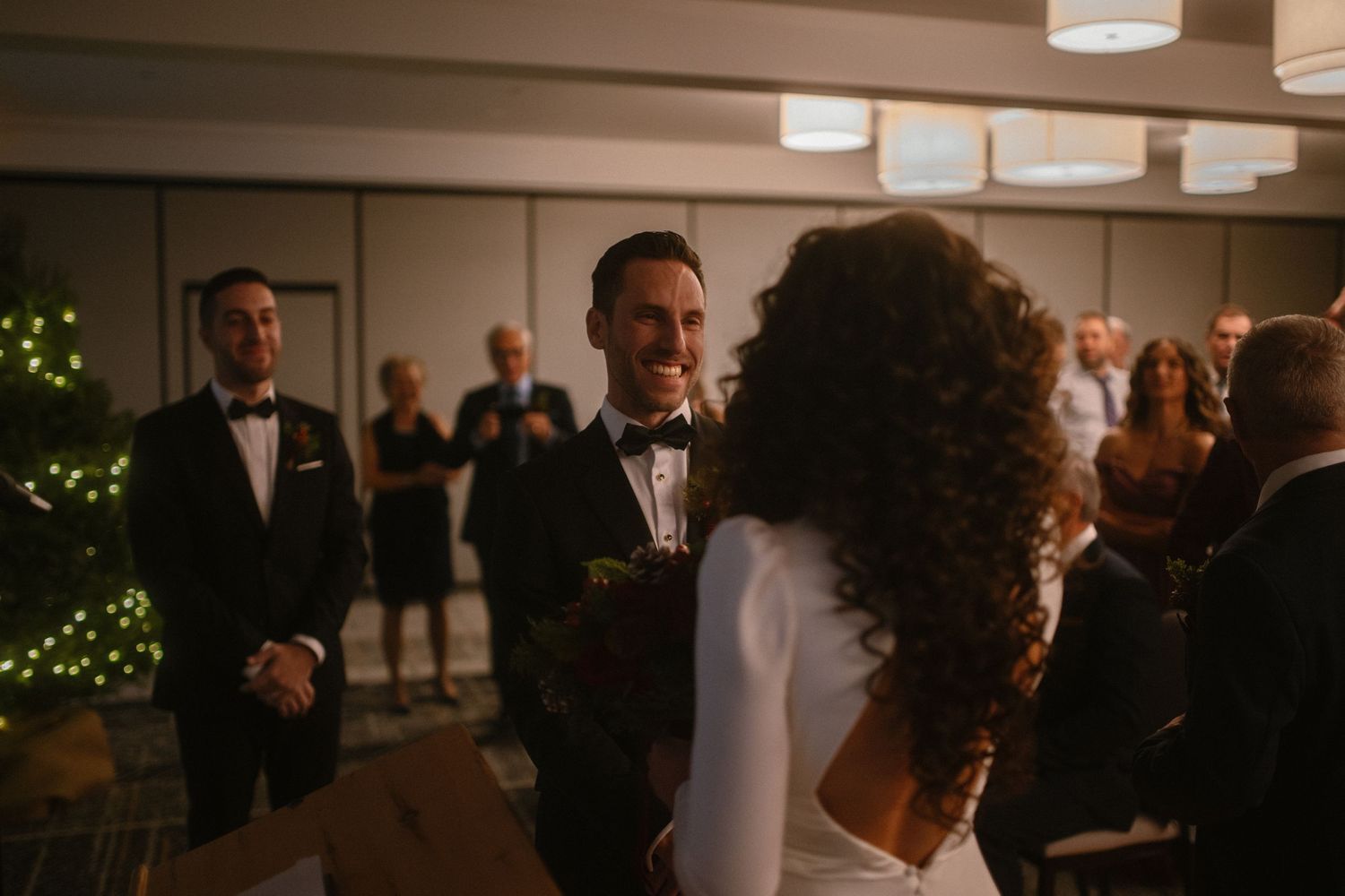 Groom smiling at bride during indoor wedding ceremony with guests and Christmas tree in background.