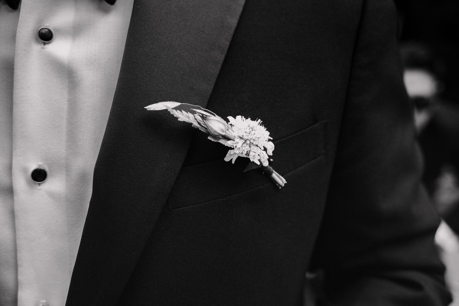 Close-up of a groom's boutonniere with white flowers and feathers pinned to a black tuxedo lapel.