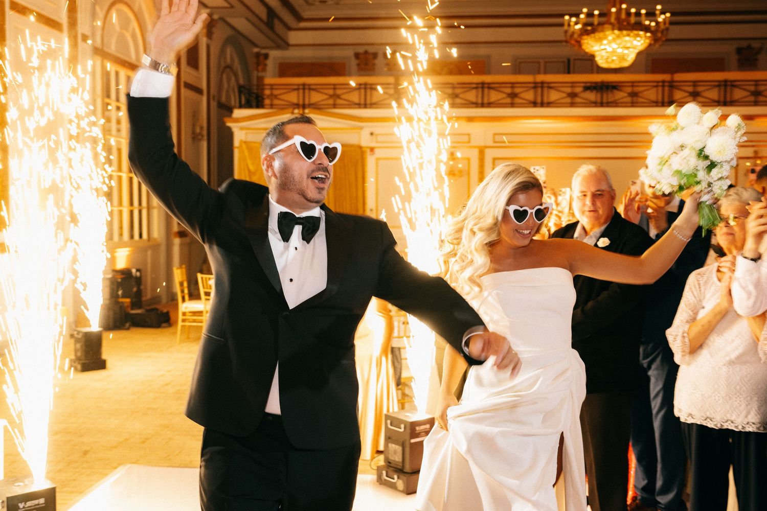 Bride and groom wearing heart sunglasses making grand entrance with sparklers at elegant wedding reception.