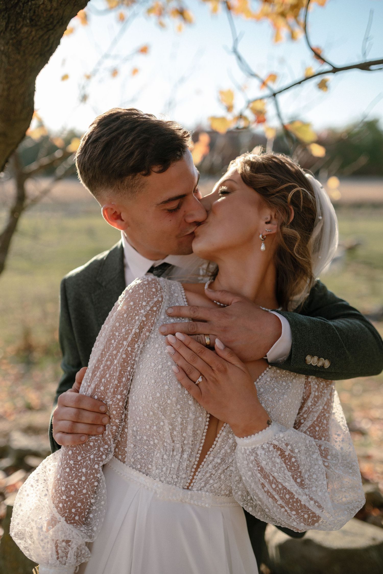 Groom kisses bride on cheek under autumn tree, bride wearing polka dot lace wedding dress.