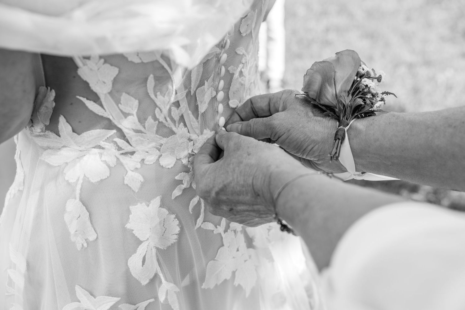 Hands fastening the back of a floral lace wedding dress in elegant black and white photography.