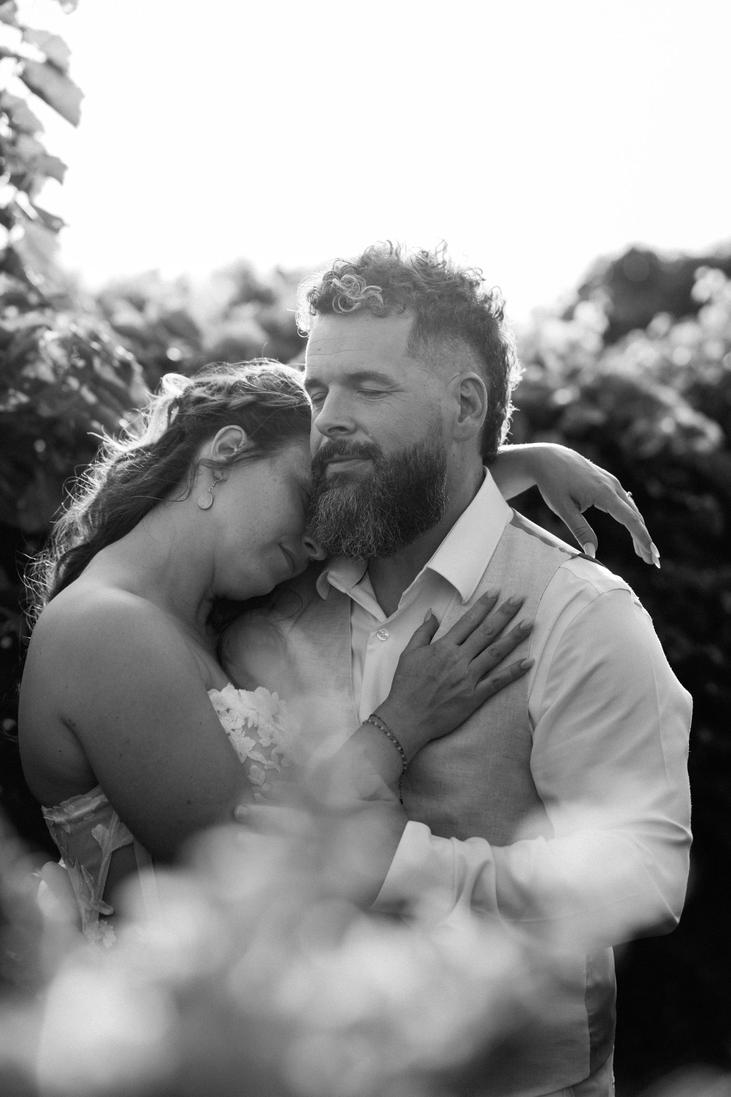 Black and white photo of a bearded groom embracing his bride outdoors, both with eyes closed in a tender moment.