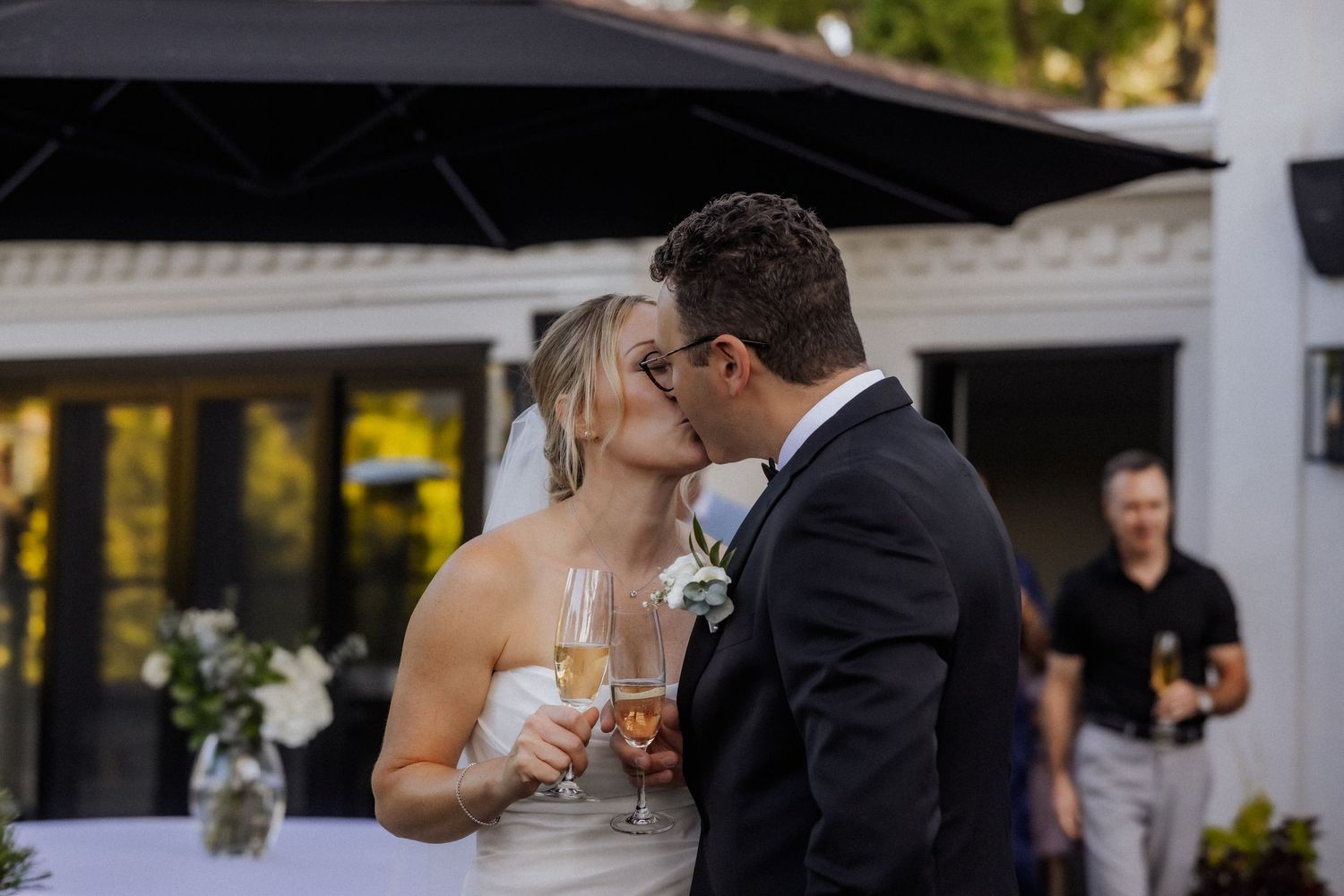 Newlywed couple kisses while holding champagne glasses during outdoor wedding reception celebration.