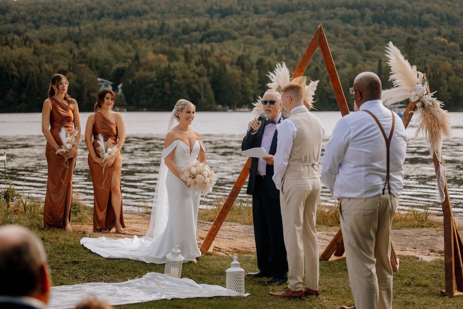 Bride and groom exchange vows at outdoor lakeside wedding ceremony with triangular pampas grass arch.