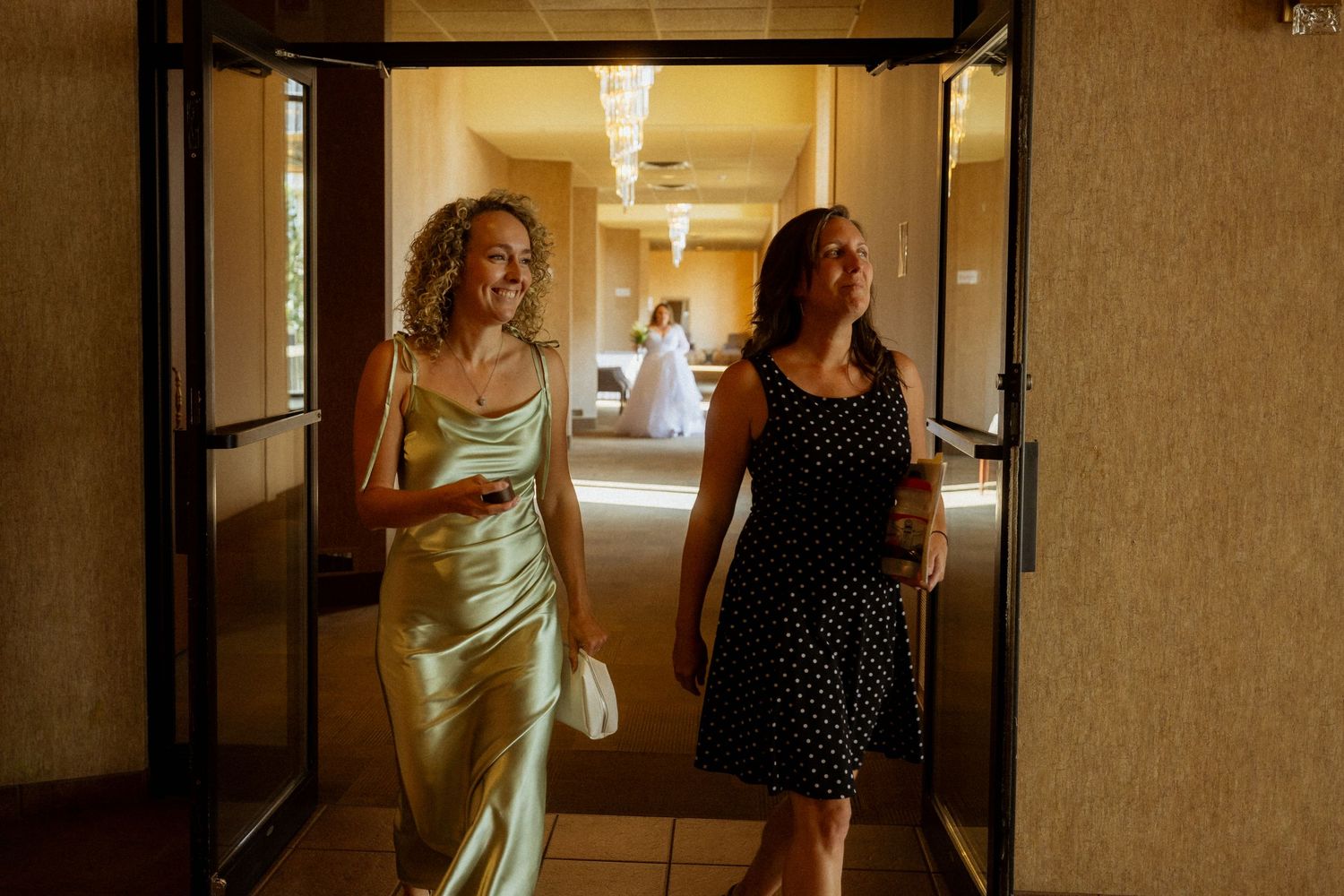 Two women walk through open doors at a wedding venue, one in a green satin dress, another in a black polka dot dress.