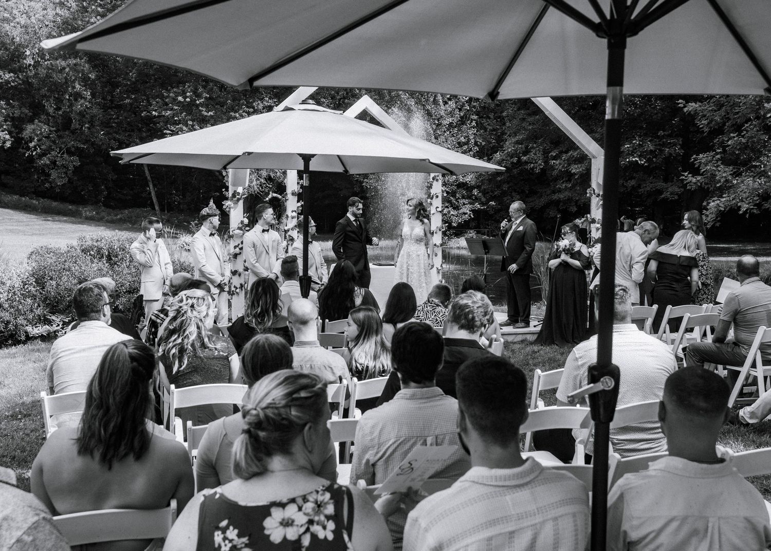 Black and white outdoor wedding ceremony with guests seated under umbrellas on a sunny day.