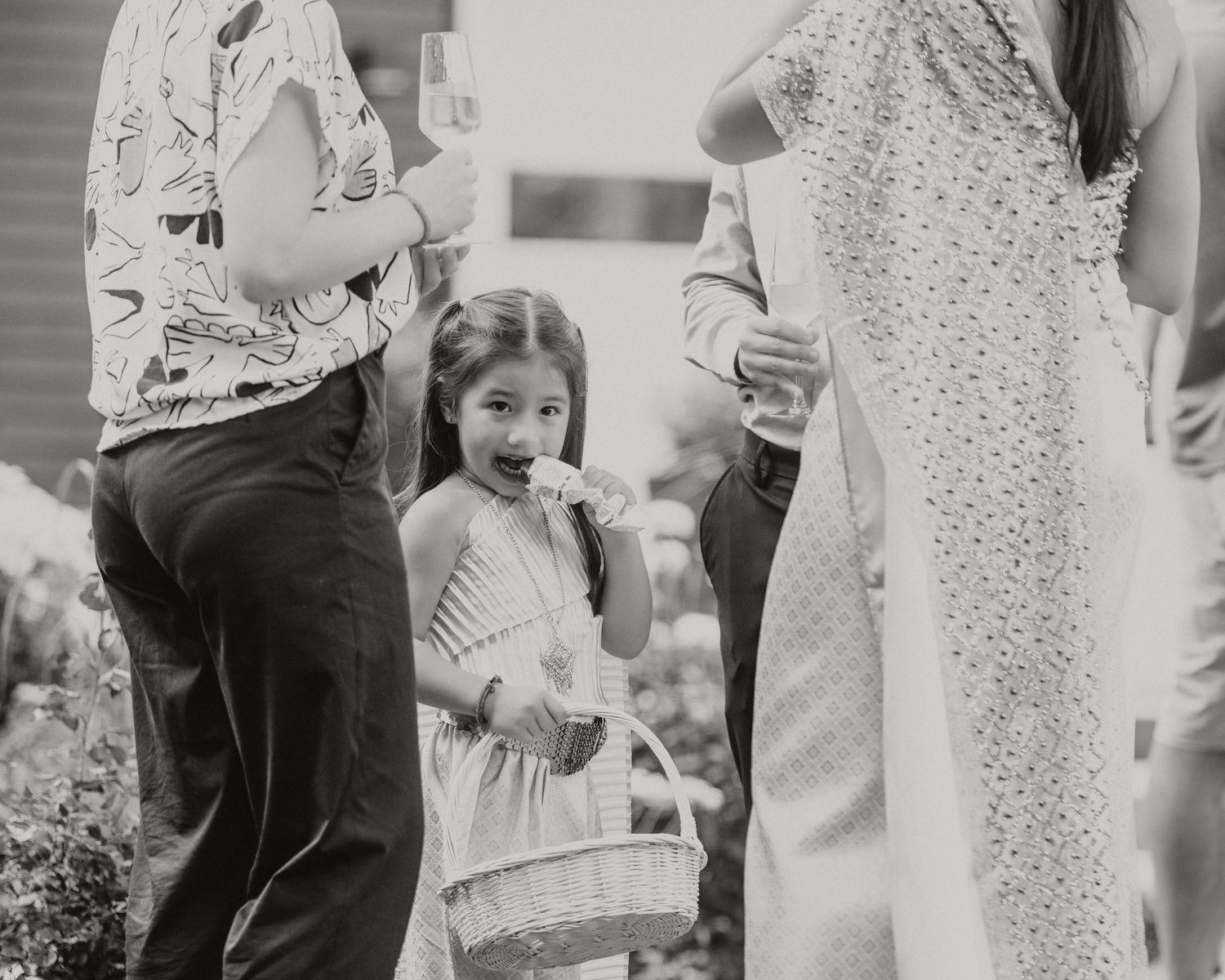 Black and white photo of a young flower girl holding a basket at an outdoor wedding ceremony.
