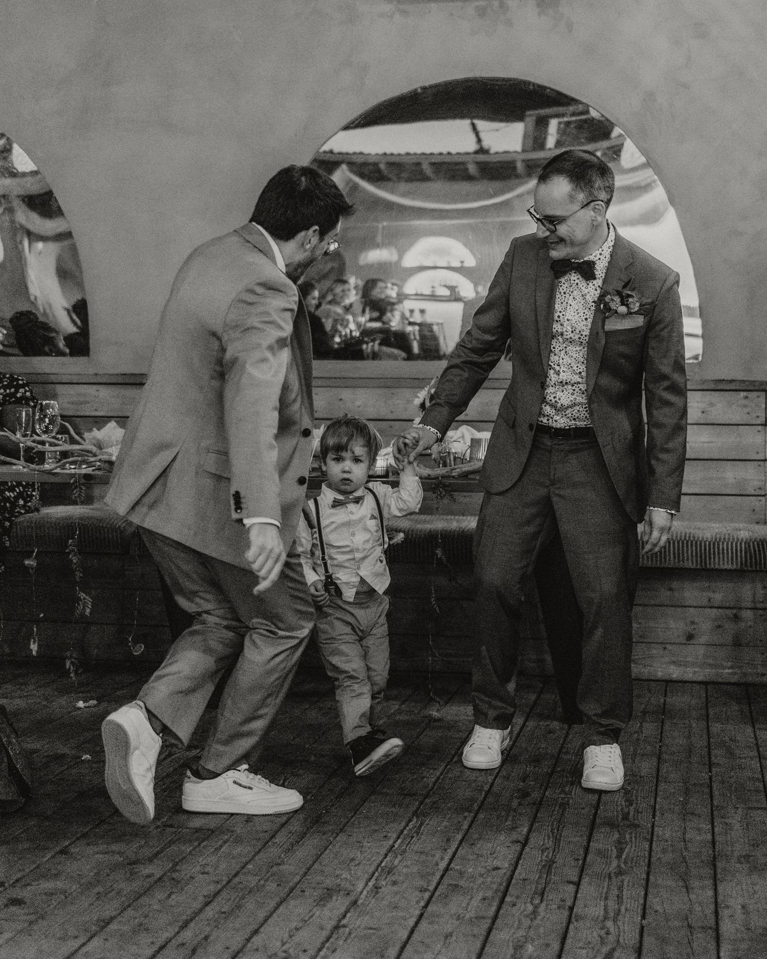 Two grooms in suits dance with a young child in suspenders at a wedding reception in black and white.