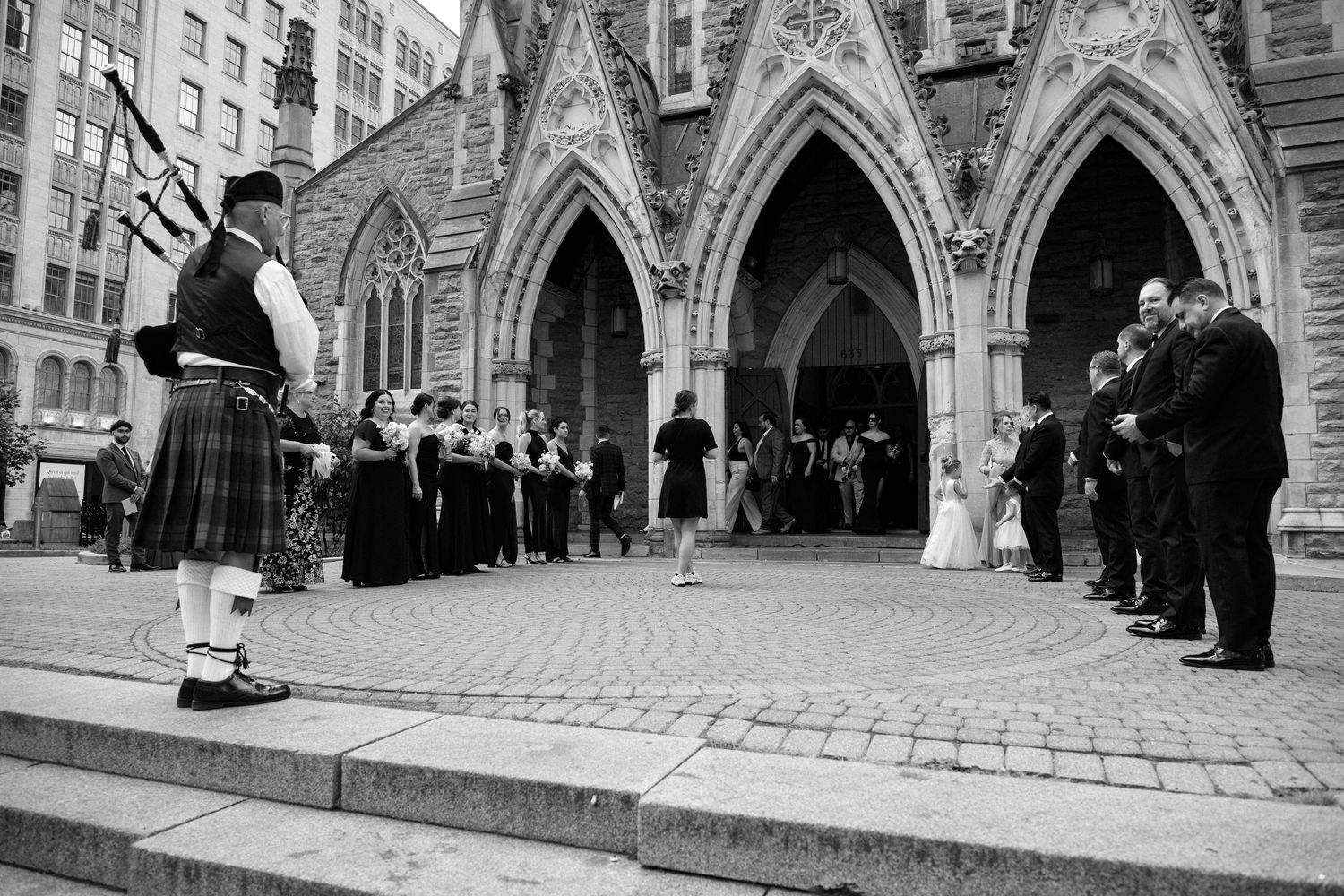 Bagpiper in tartan kilt performing outside gothic stone church entrance for wedding guests in black and white.