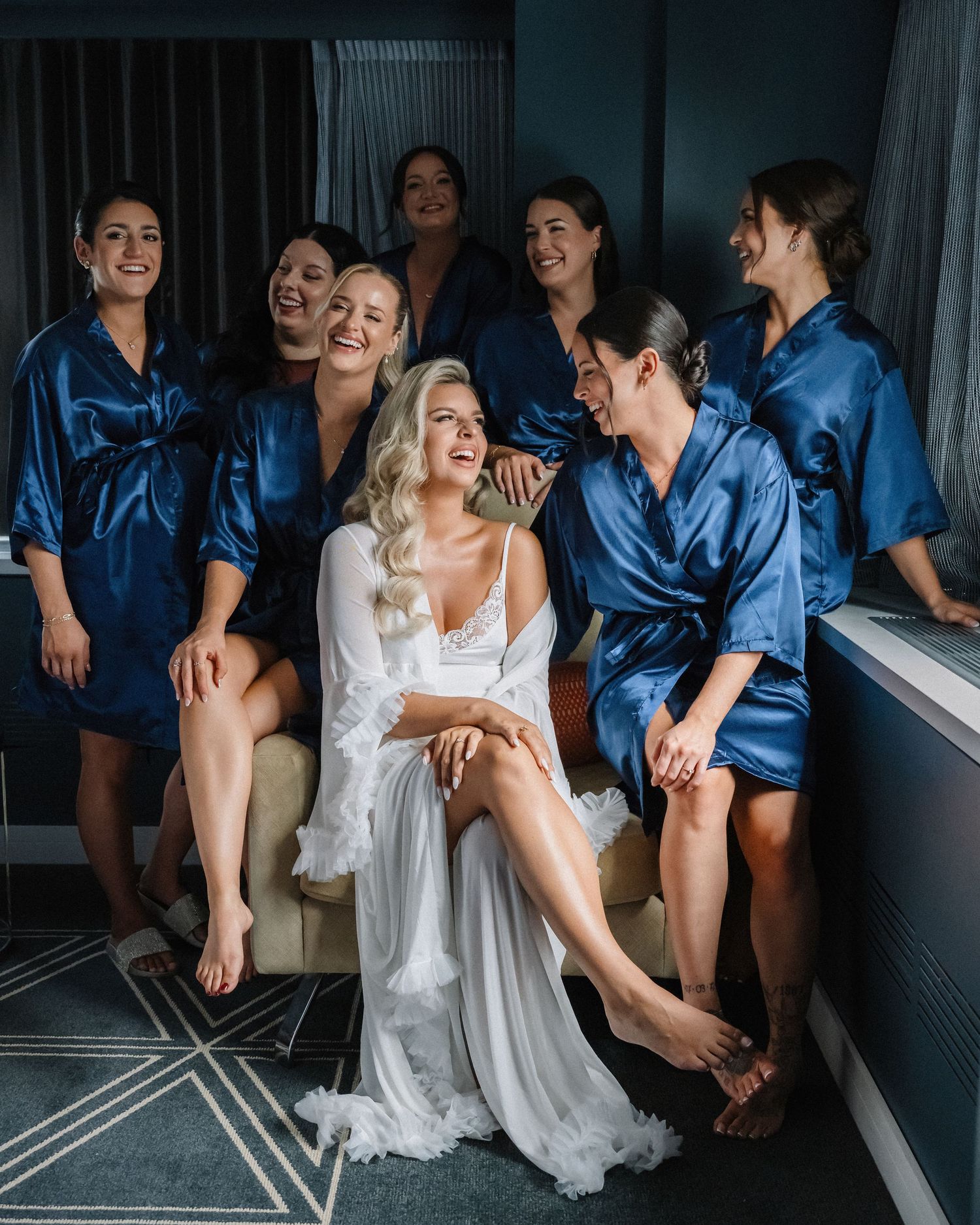 Bride in white lace robe laughing with bridesmaids in blue satin robes during wedding preparations.