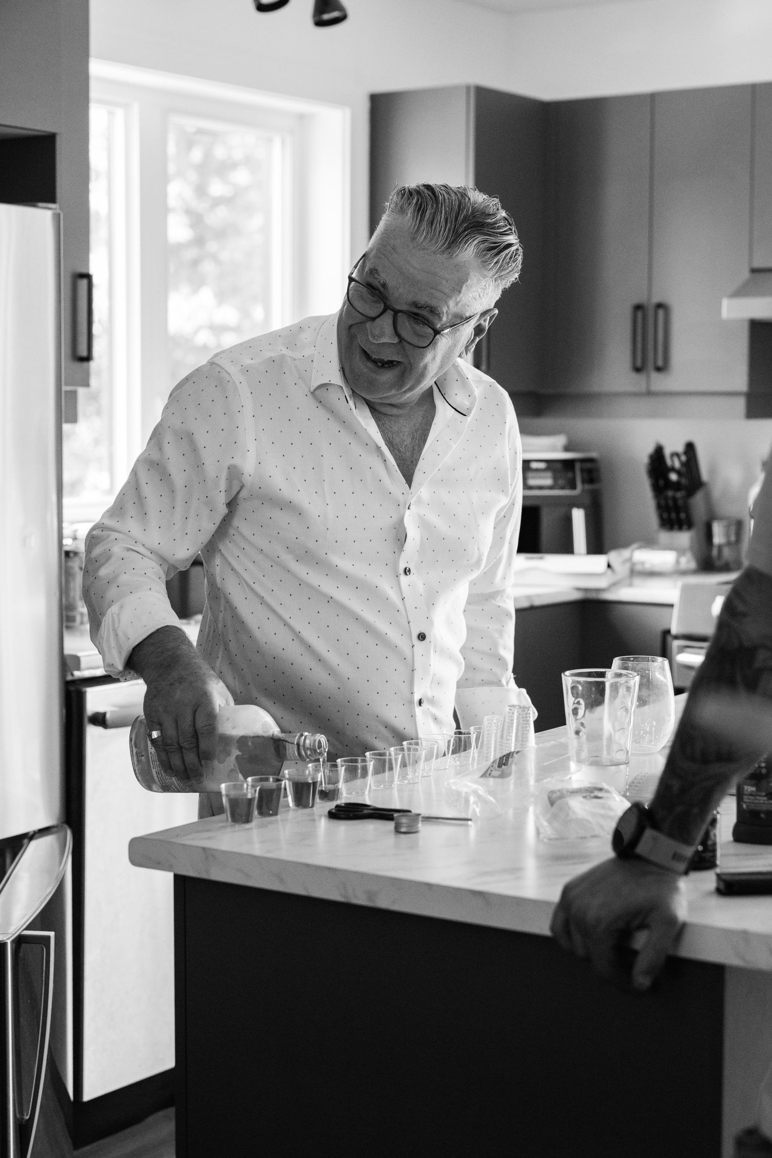 Man in white shirt pouring shots in a modern kitchen at a wedding celebration in black and white.