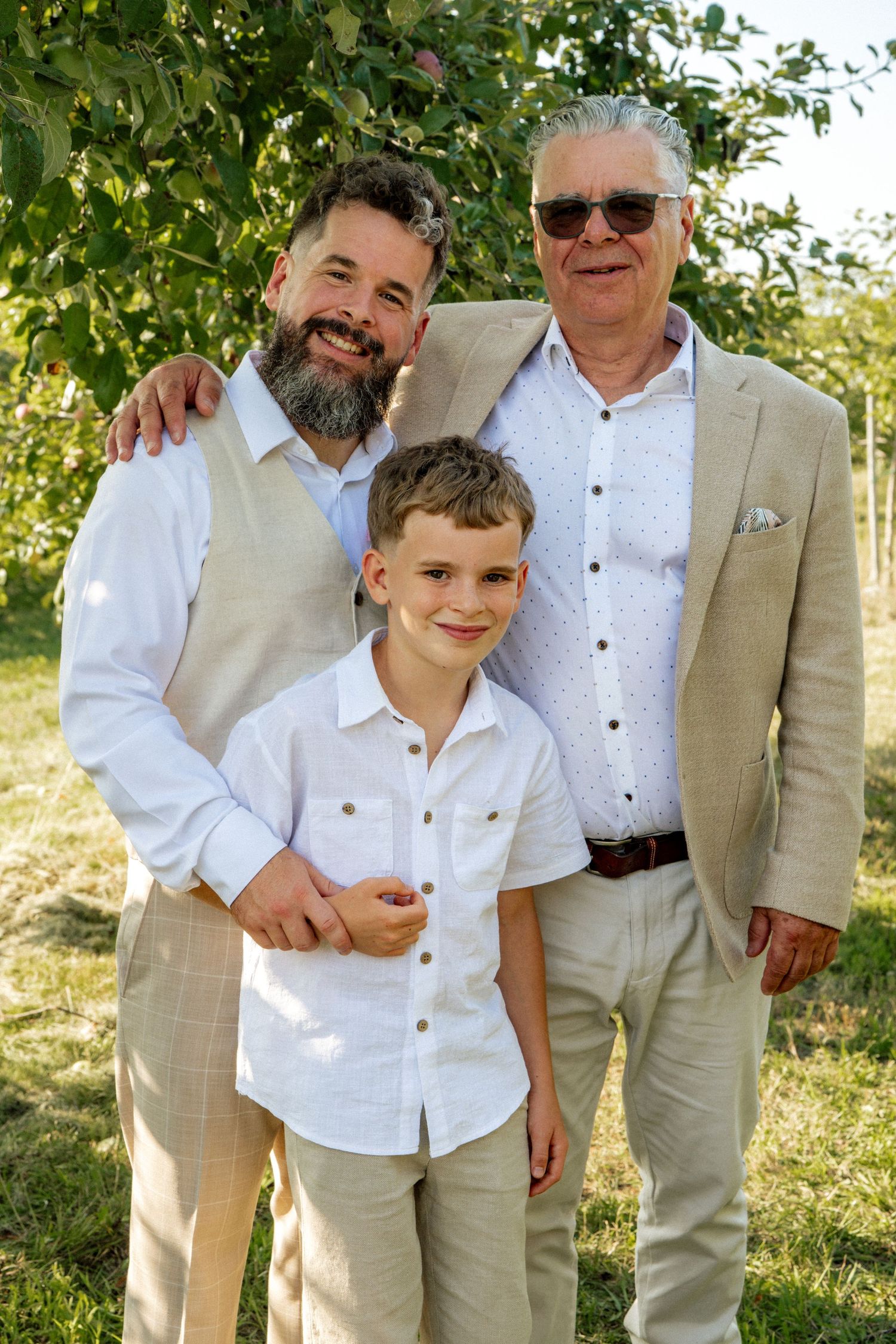 Three generations of men in beige suits pose together outdoors at a wedding celebration.
