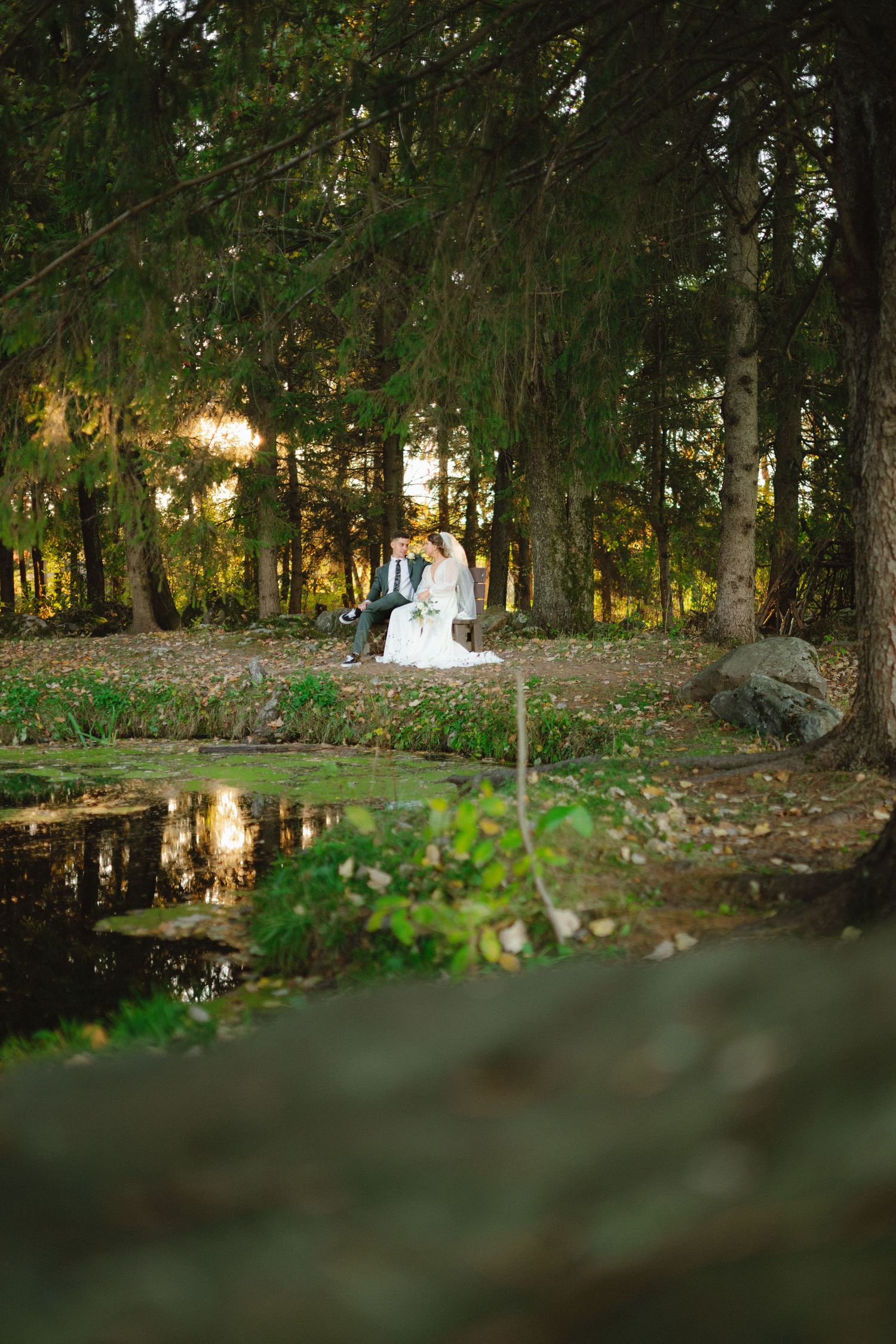 Bride and groom sitting together near a peaceful pond surrounded by lush green trees.
