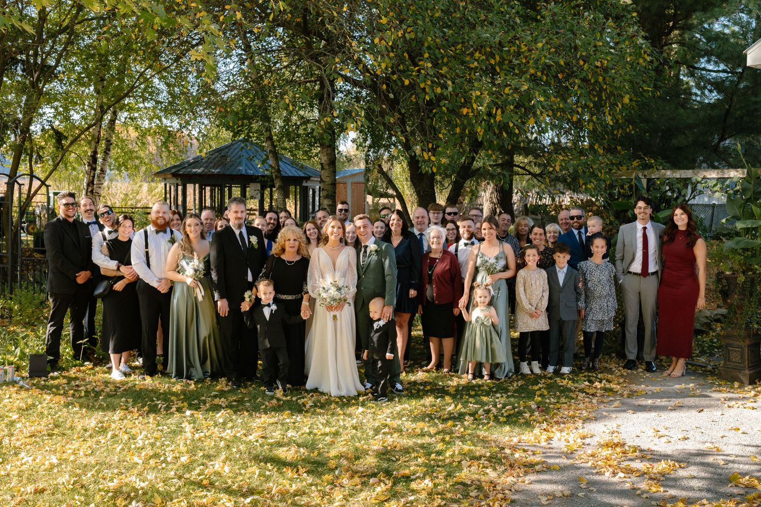 Large wedding party group photo surrounded by autumn foliage near a gazebo in a park setting.