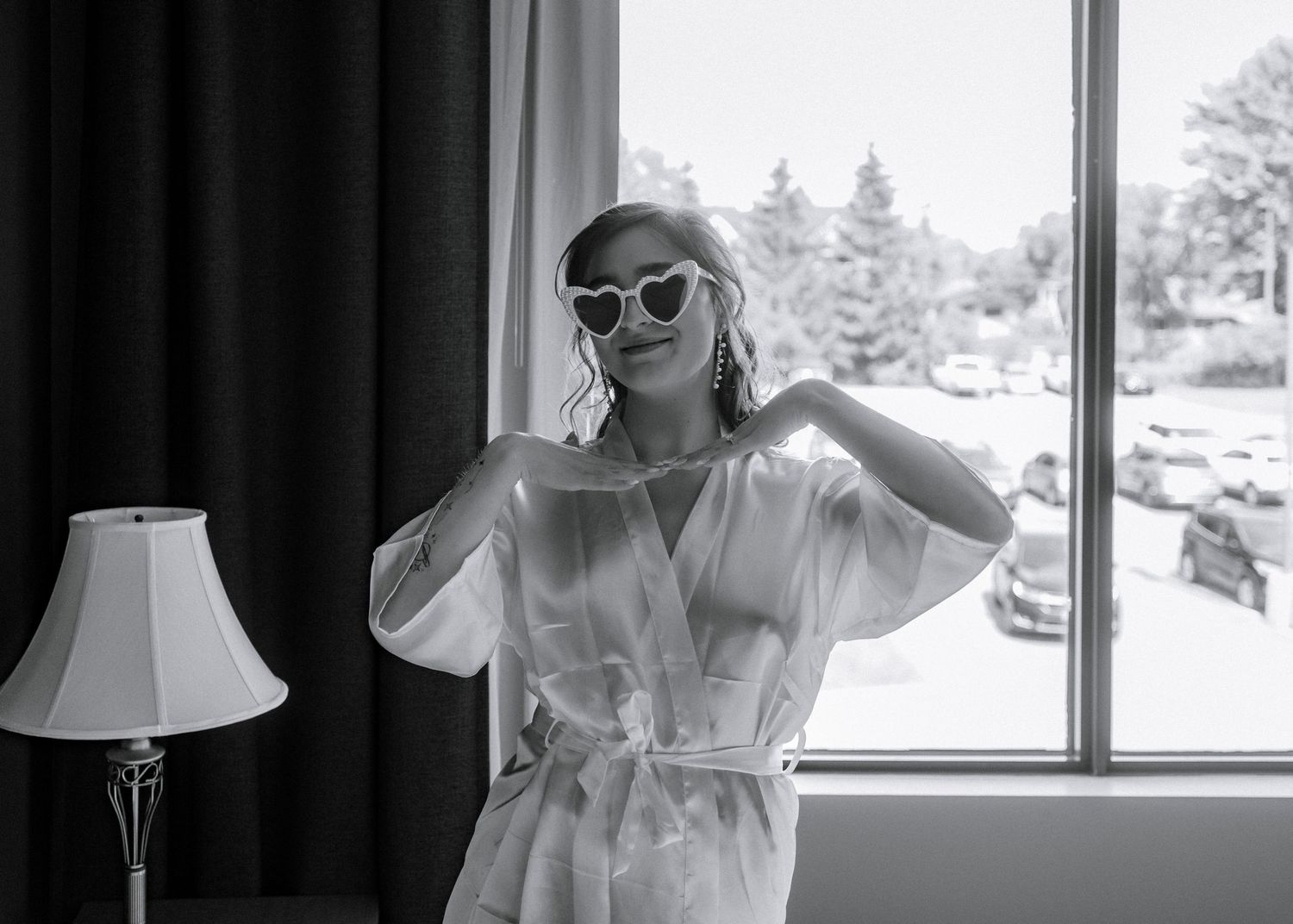 Bride in white robe and heart-shaped sunglasses posing by window in black and white photo.