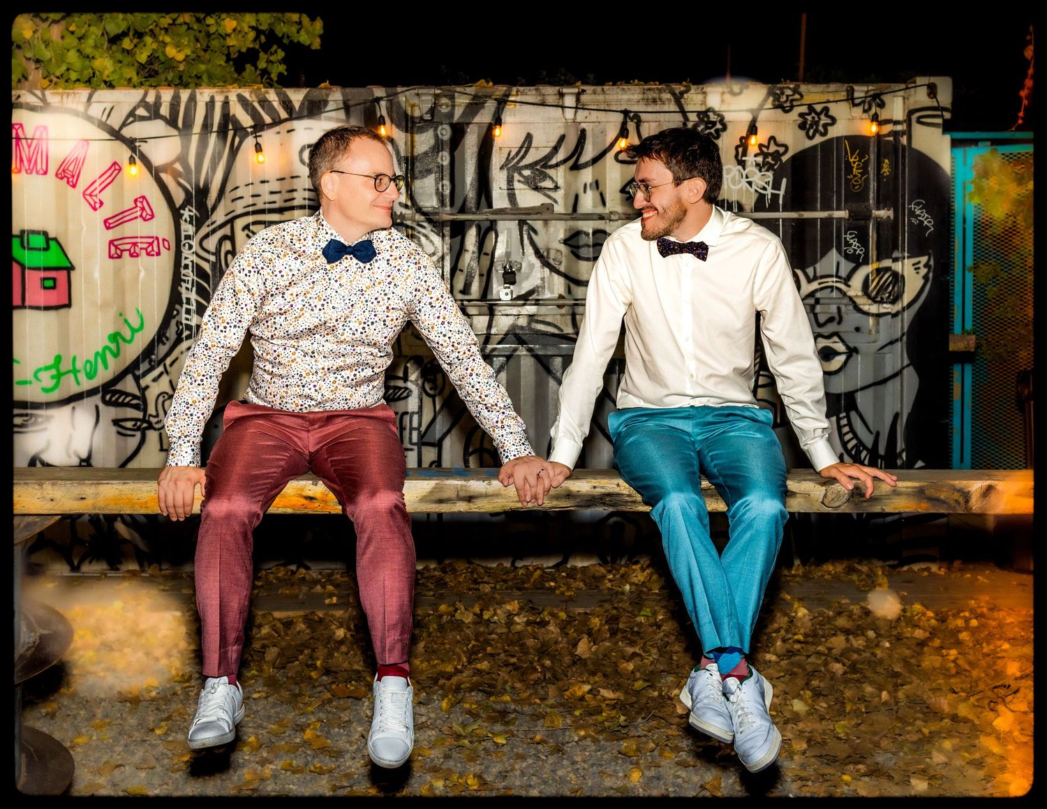 Two grooms in bow ties hold hands on a bench in front of a colorful graffiti wall.