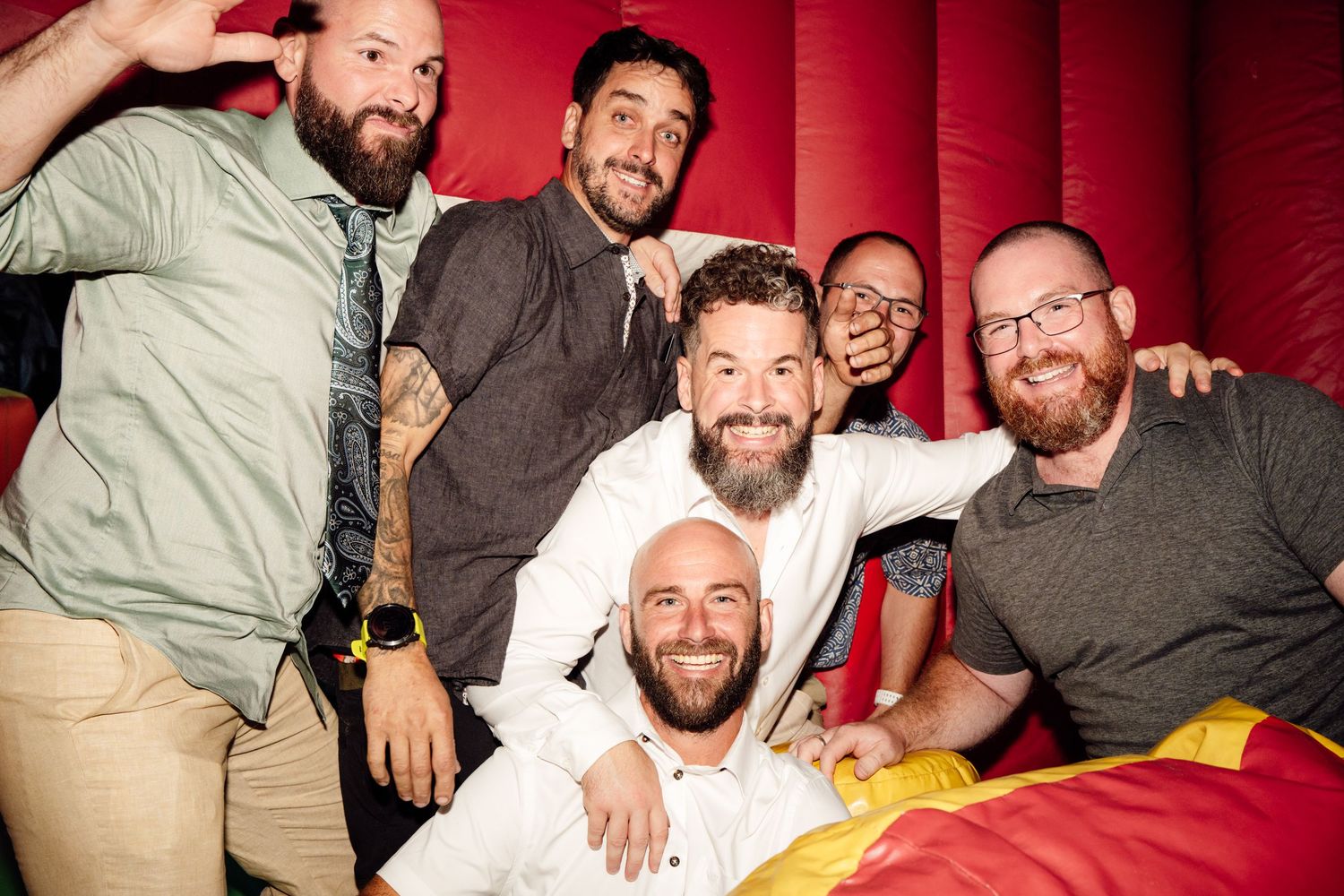 Group of six smiling groomsmen pose together on a colorful inflatable bouncy castle at reception.