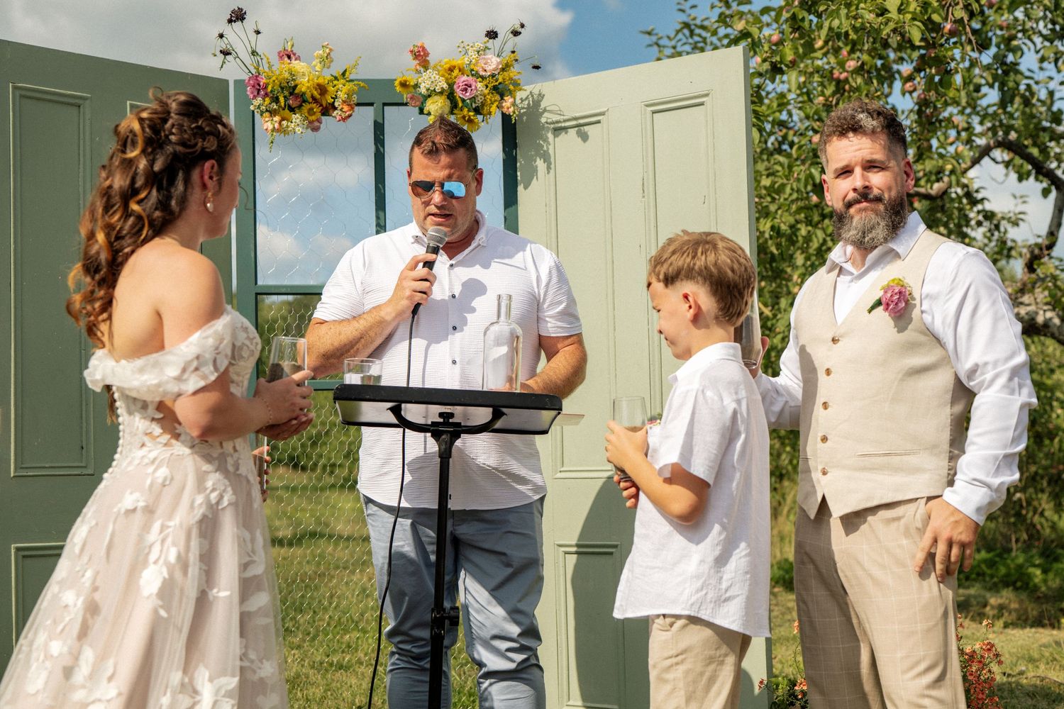 Wedding ceremony outdoors with officiant, groom, and child standing before green doors altar.
