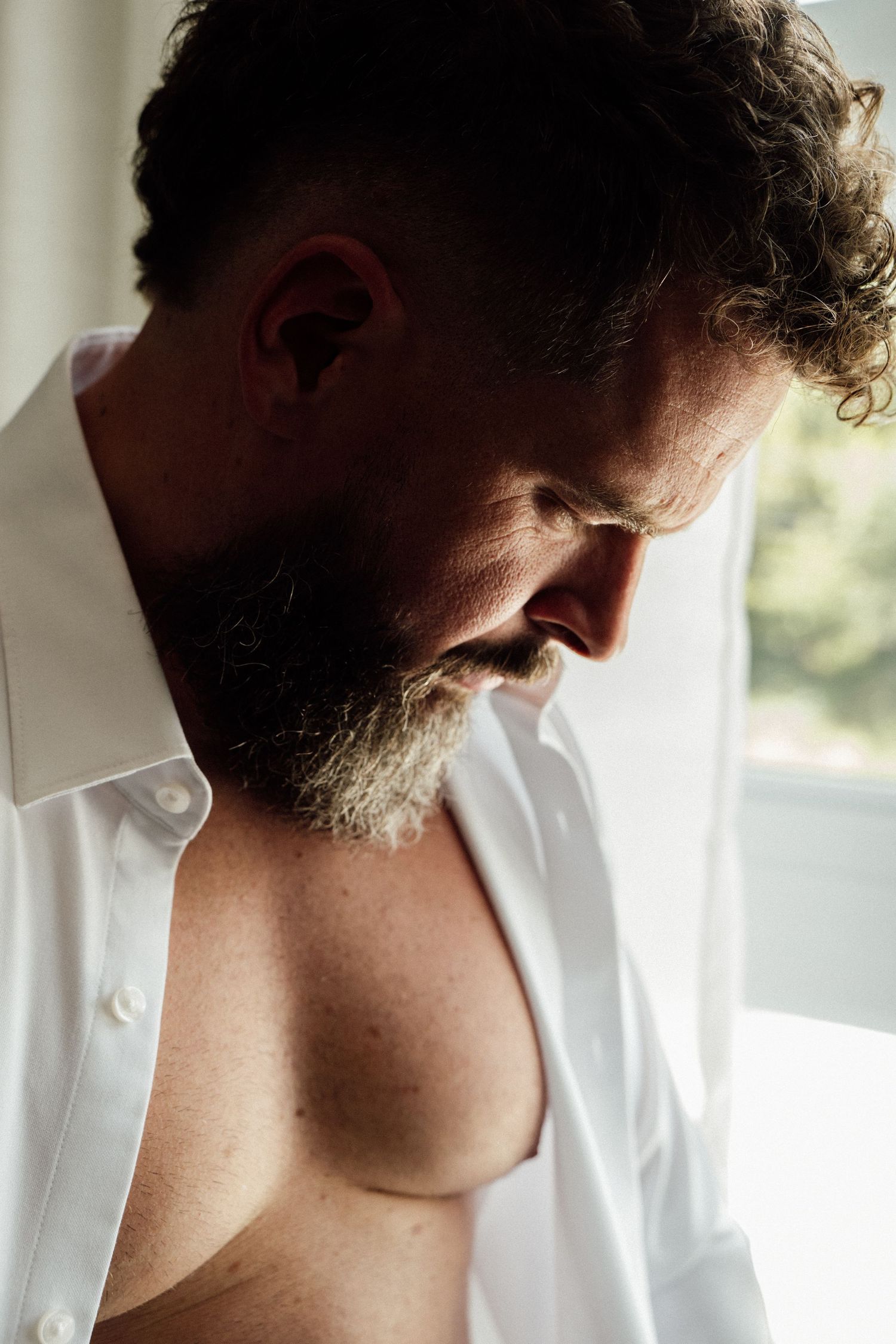 Bearded groom in open white shirt getting ready near window on wedding day.