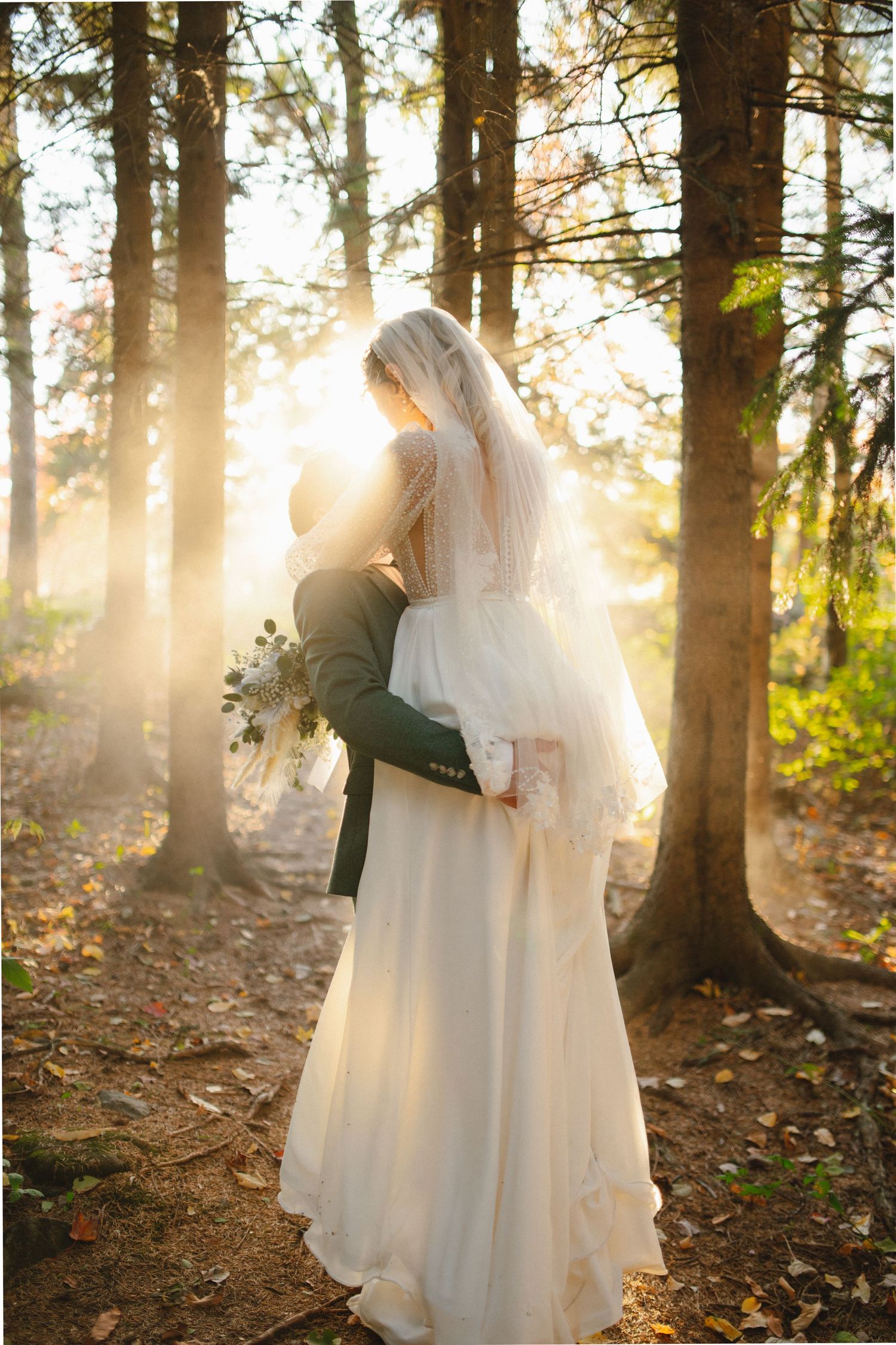 Bride in flowing white gown and veil embraces groom in green suit among sunlit trees in an autumn forest.