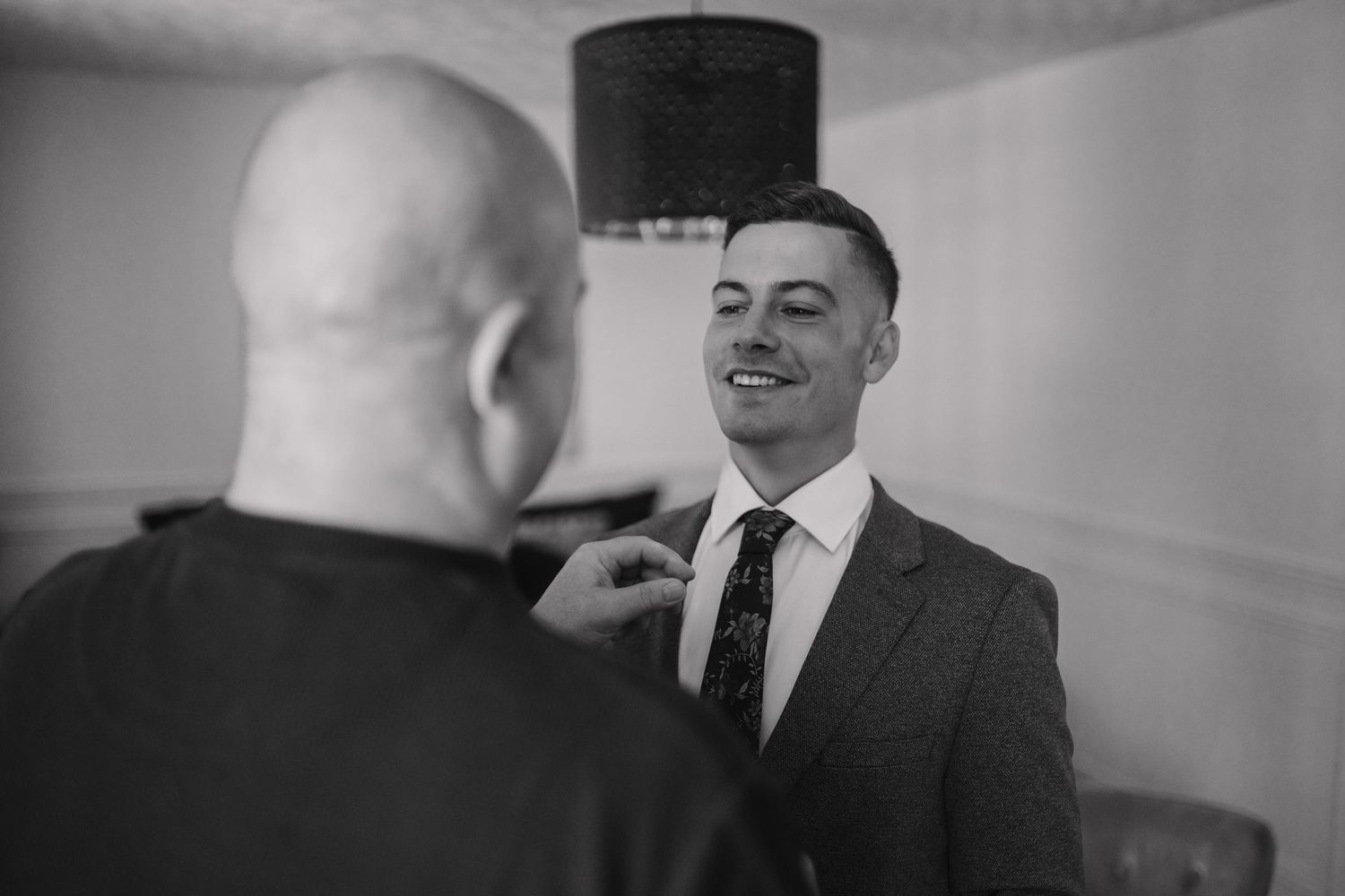 Black and white photo of a groom smiling as a man adjusts his tie before a wedding ceremony.