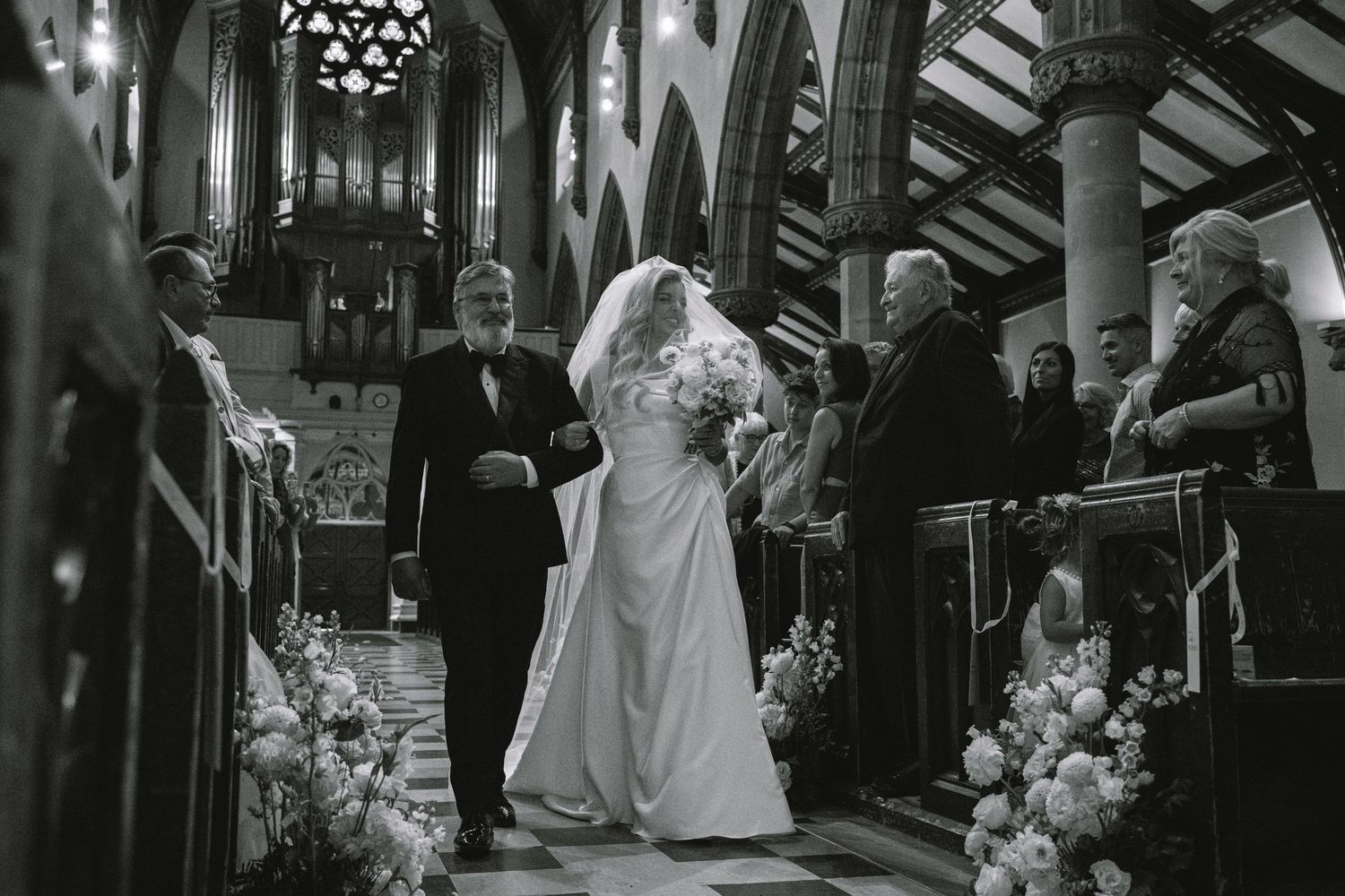 Bride walks down the church aisle with her escort past floral arrangements and wedding guests.