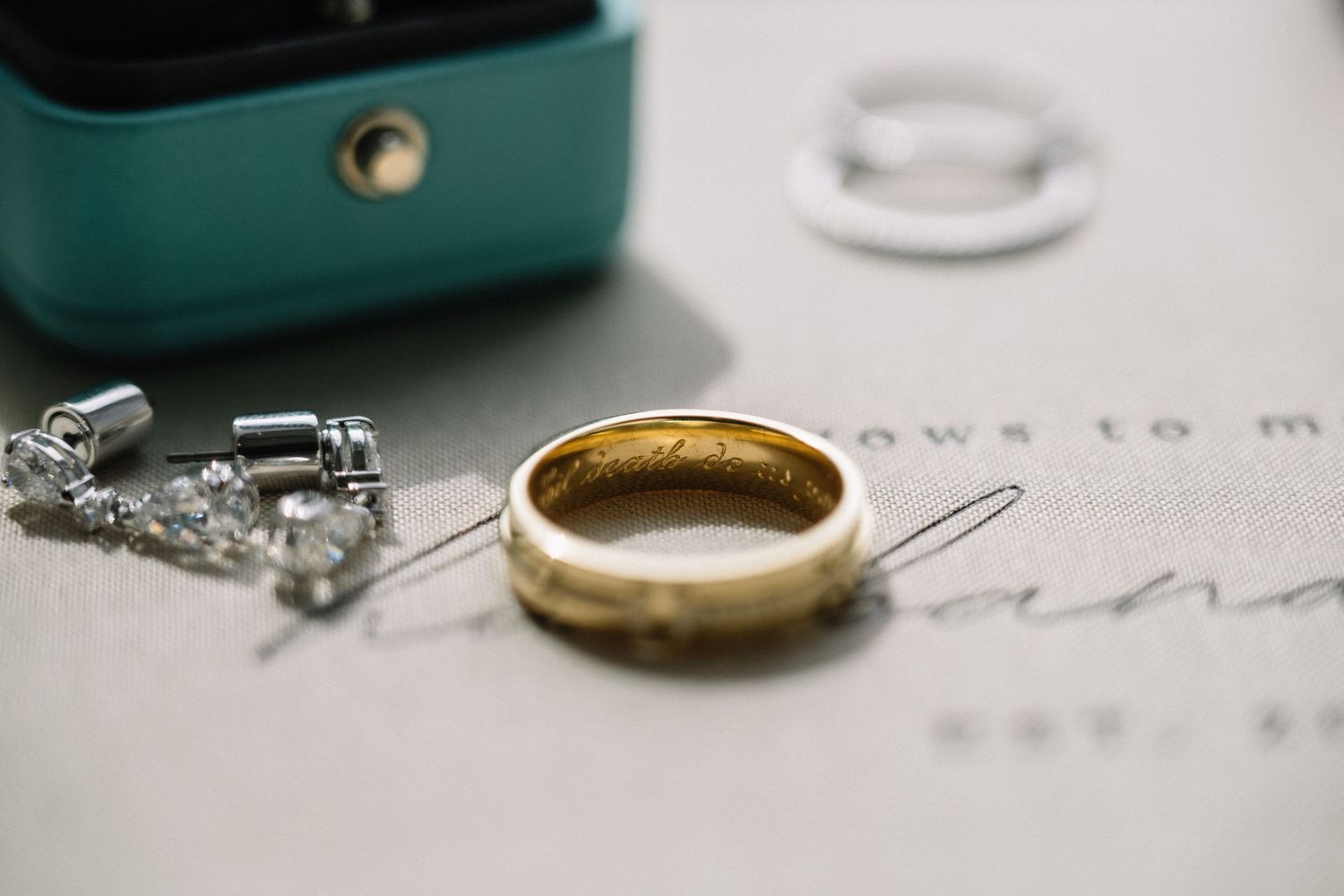 Gold wedding band with engraving beside diamond earrings and a Tiffany blue ring box on elegant stationery.