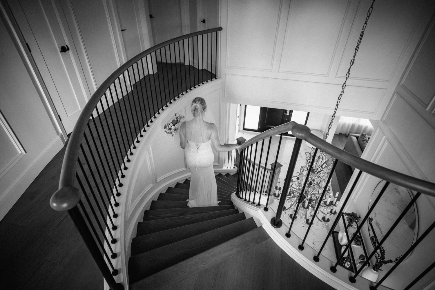 Elegant bride in white gown descends grand curved staircase in black and white wedding portrait.