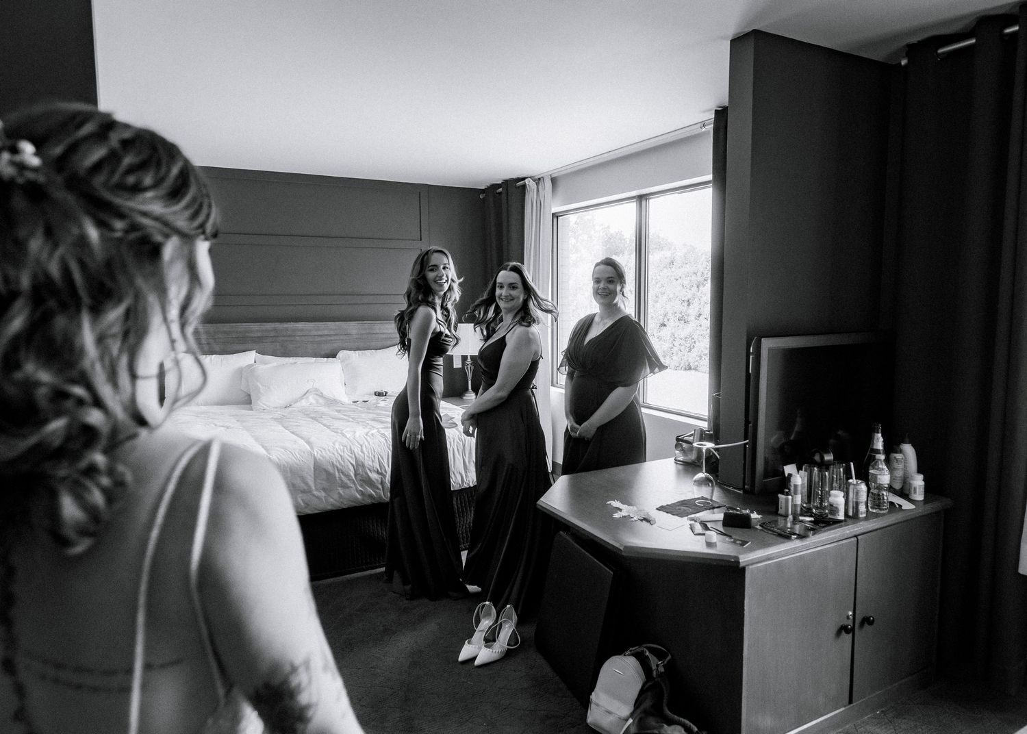 Black and white photo of bridesmaids reacting with joy as bride reveals her wedding dress in a hotel room.