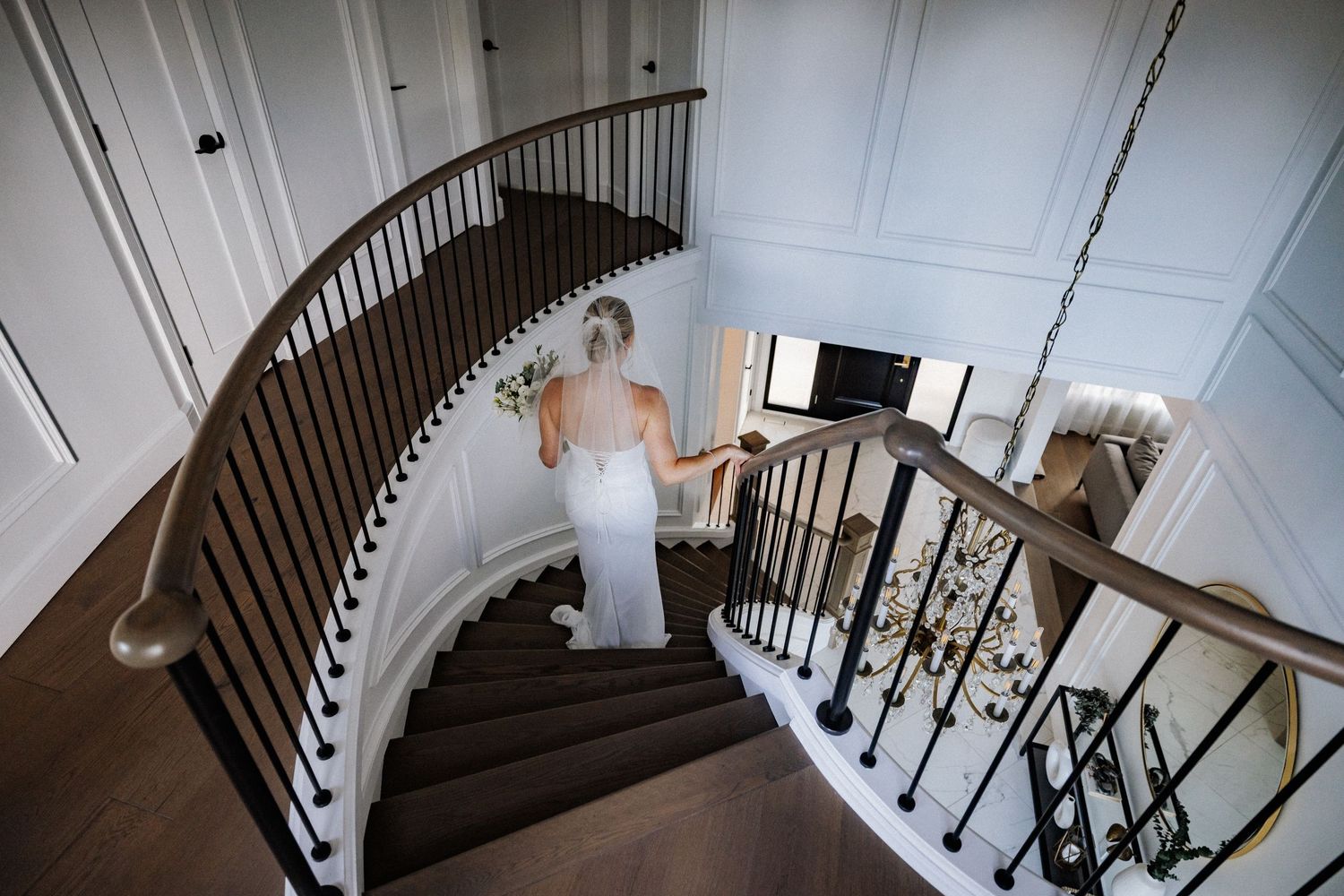 Bride in white gown descending grand curved staircase with ornate iron railing.