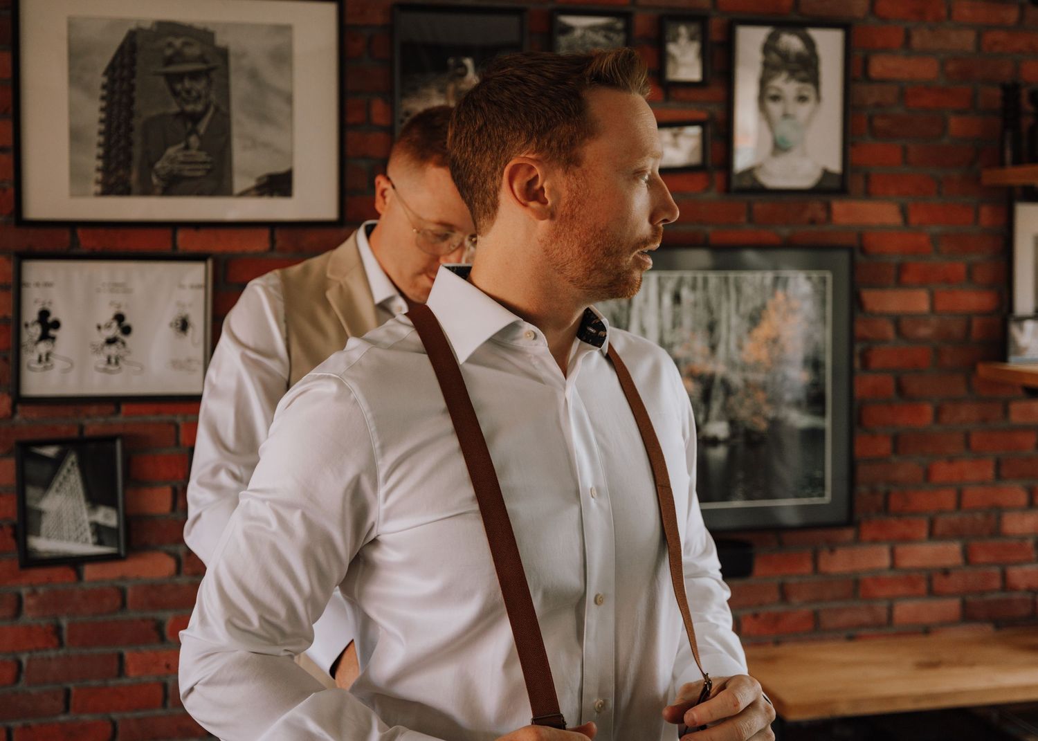 Two men in white dress shirts with brown suspenders standing in a rustic brick-walled room with framed photos.