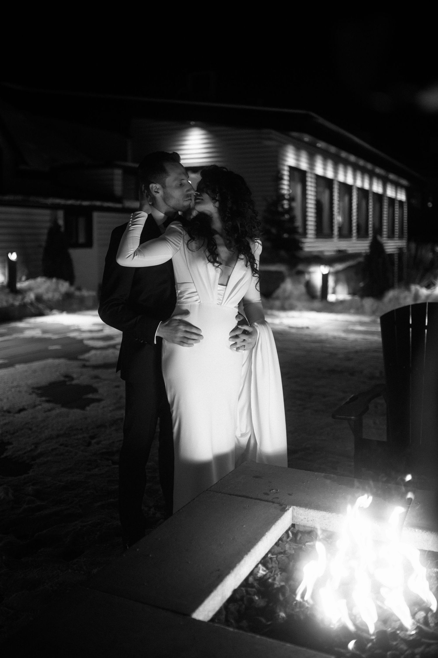 Newlyweds share romantic kiss outdoors at night beside glowing fire pit in black and white photo.