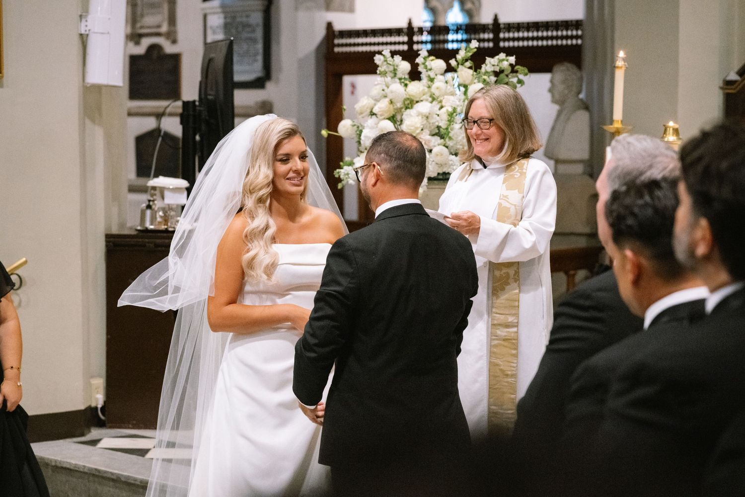 Smiling bride and groom exchanging vows at a church wedding ceremony with an officiant.