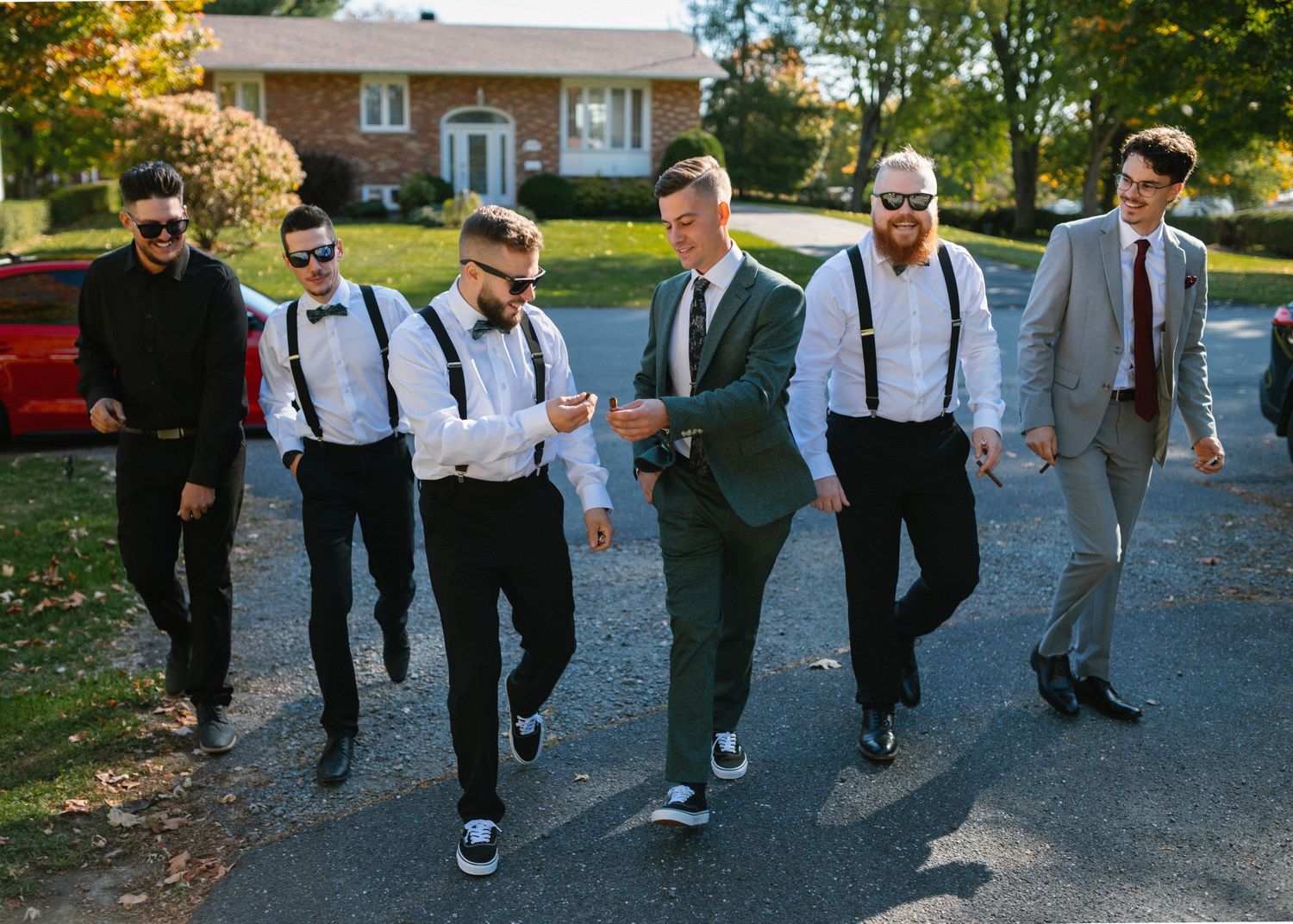 Groom and groomsmen in suspenders and bow ties walking outdoors on a driveway before a wedding ceremony.