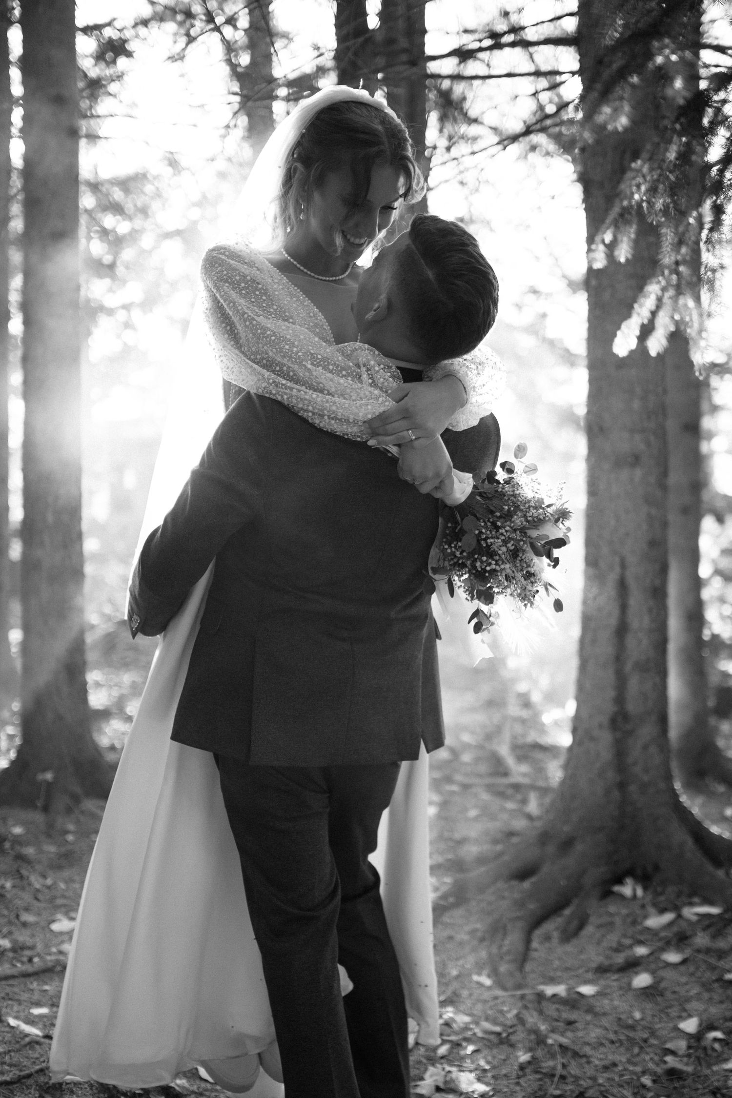 Black and white photo of groom lifting bride in forest with sunbeams shining through trees.