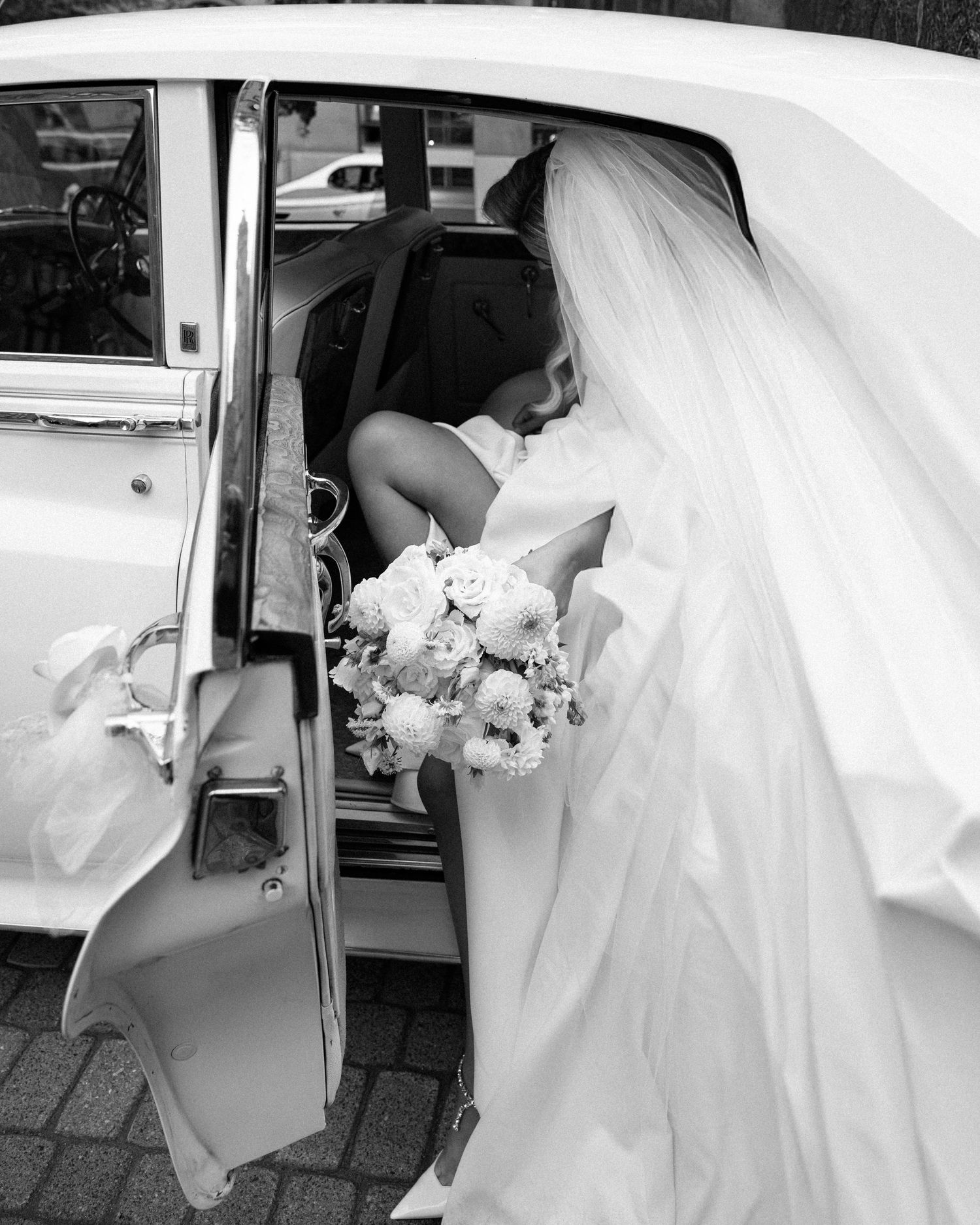 Black and white photo of bride in veil holding bouquet entering vintage white car.