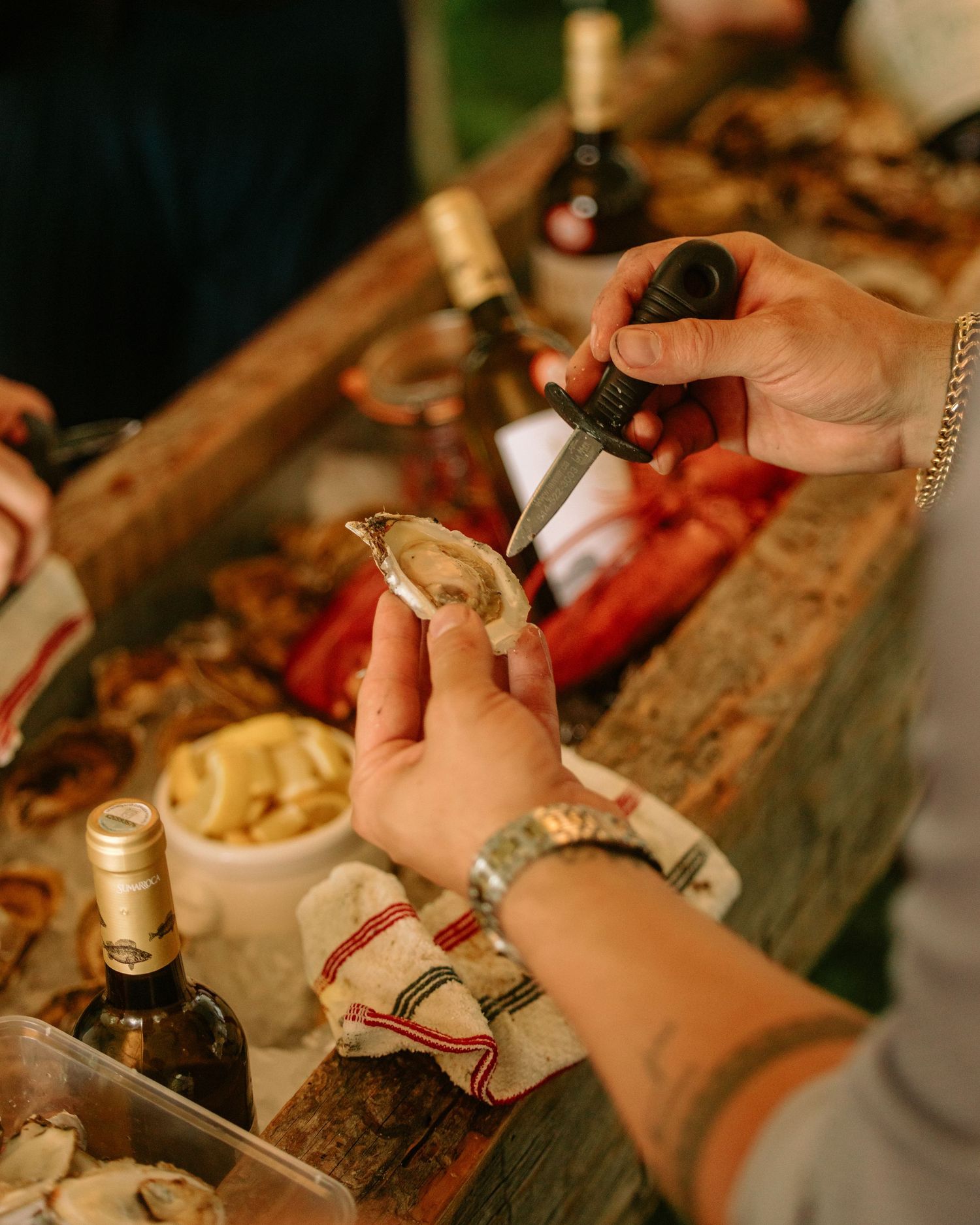 A person shucking a fresh oyster with a knife at an outdoor seafood event with wine bottles nearby.