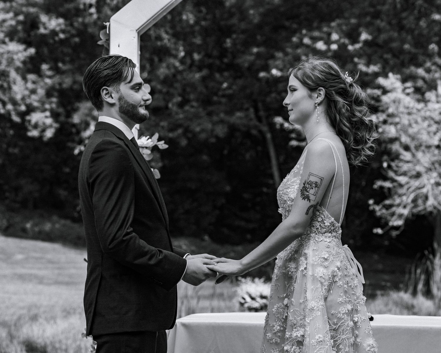 Black and white photo of bride and groom holding hands at outdoor wedding ceremony beneath a white arch.
