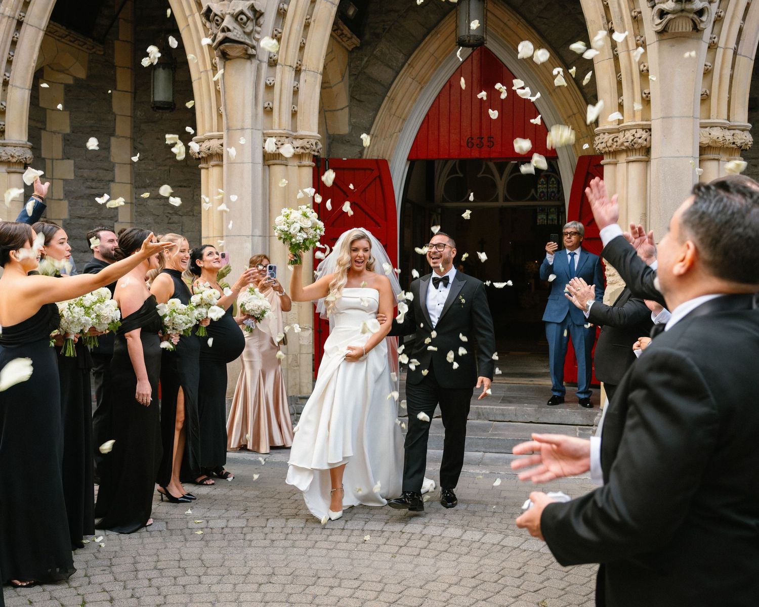 Bride and groom exit church amid rose petal toss, flanked by bridesmaids in black gowns and groomsmen.