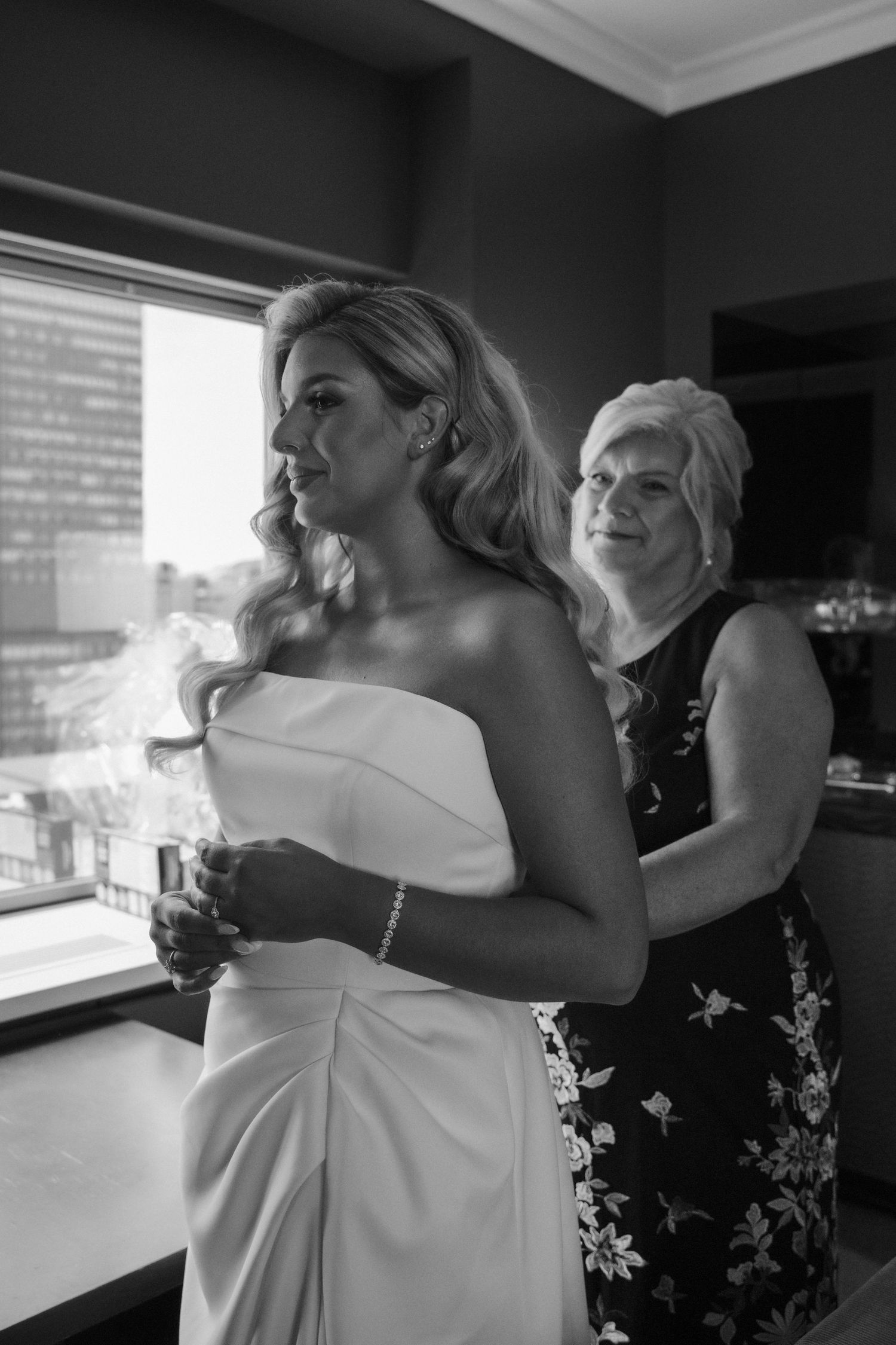 Black and white photo of mother helping bride into strapless wedding gown by a window.