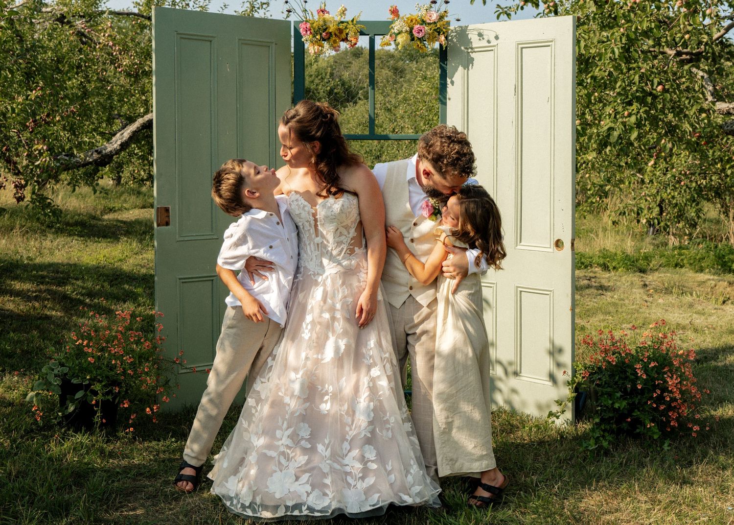 Bride and groom embrace with two young children in lush garden wedding ceremony near vintage doors.