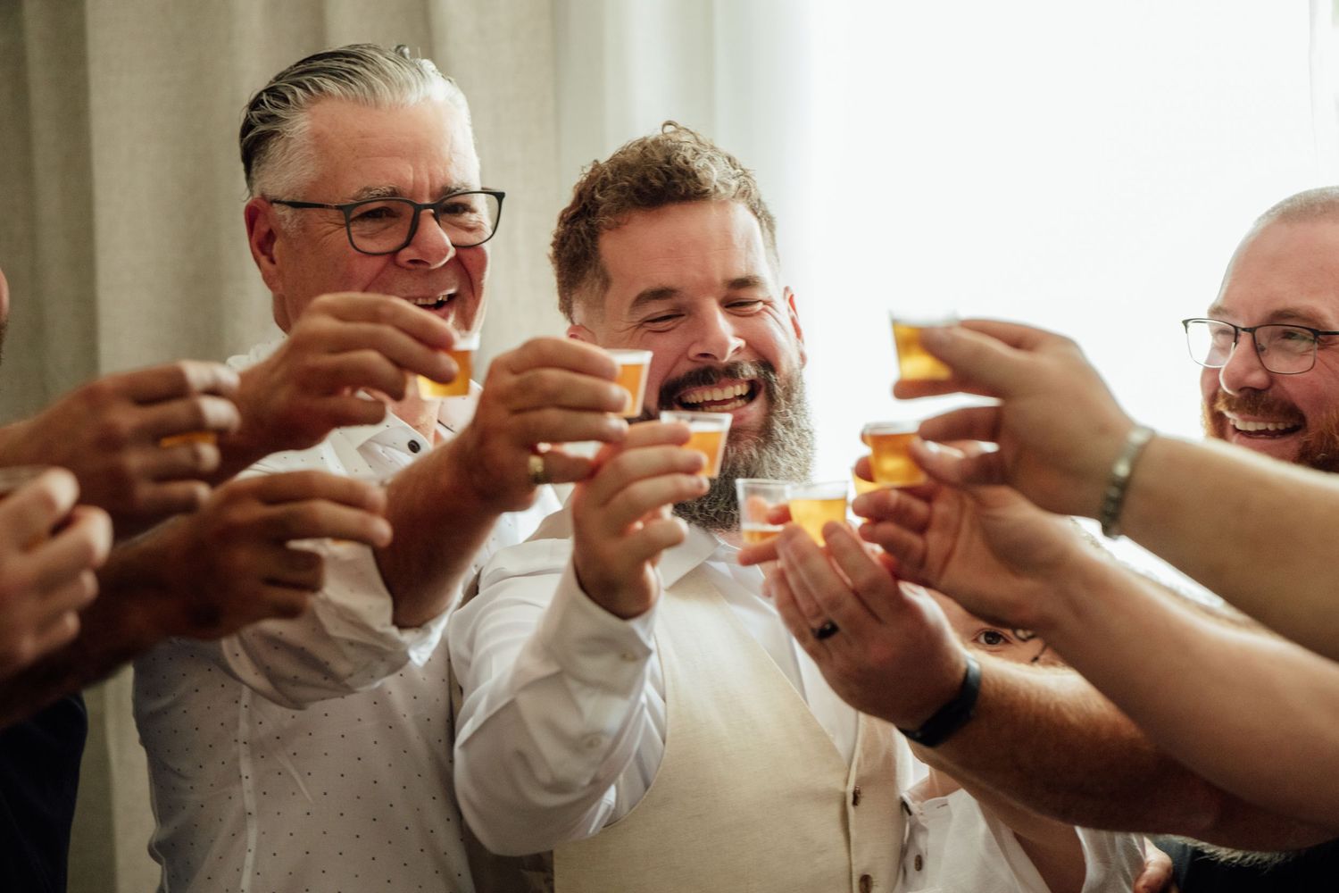 Group of men laughing and toasting with drinks at a celebration or wedding event.
