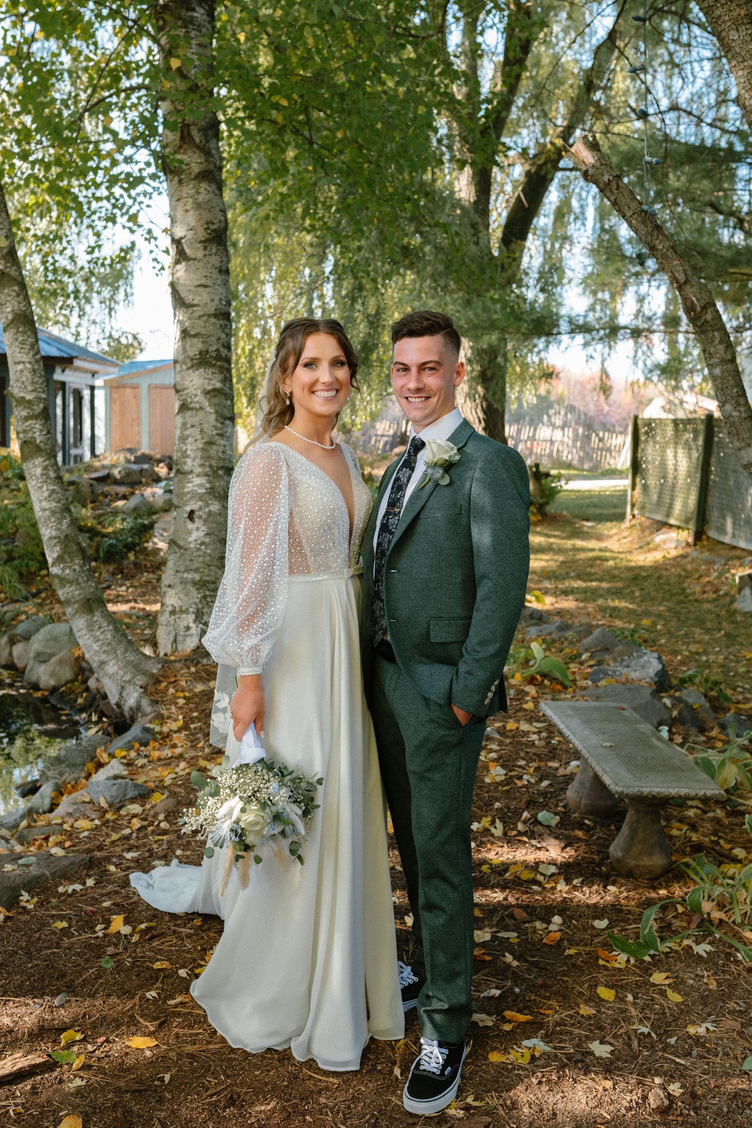 Bride in white lace gown holds bouquet beside groom in green suit among birch trees in autumn garden.