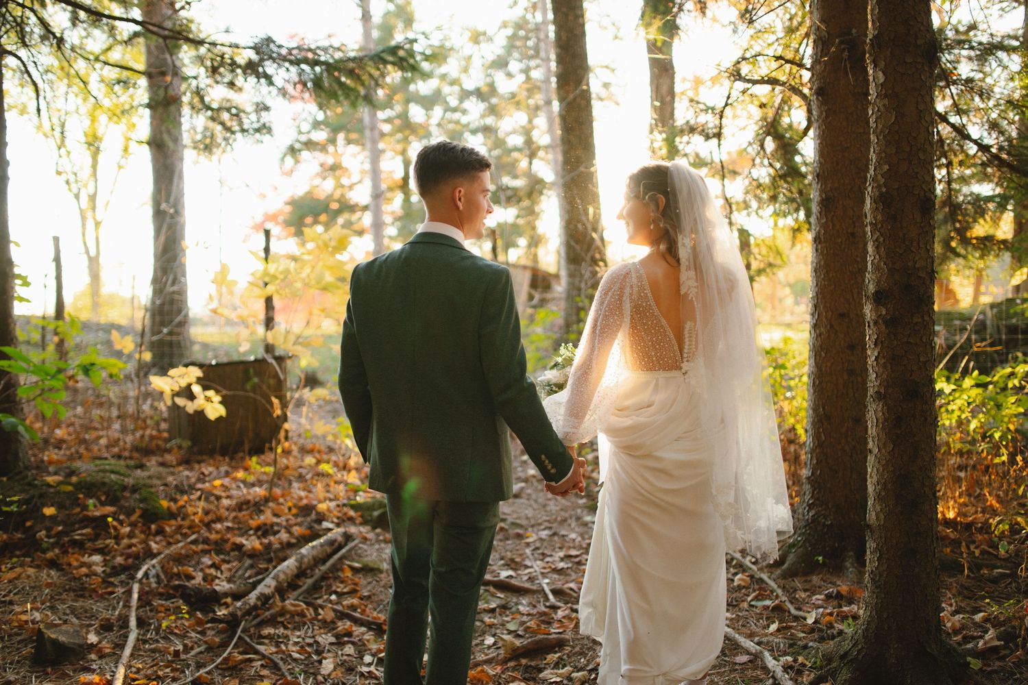Bride and groom walk hand in hand through a sunlit autumn forest after their wedding.