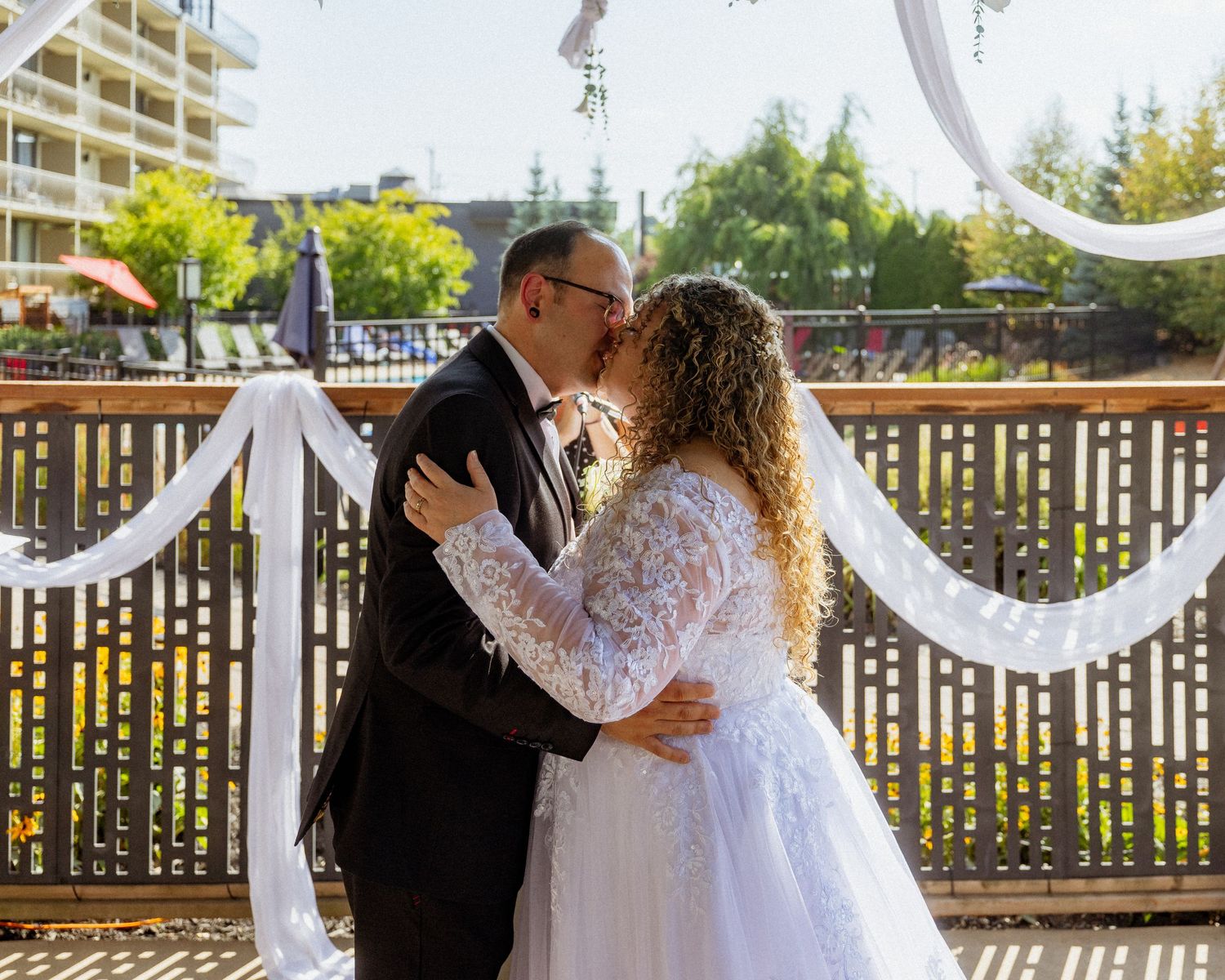 Bride and groom share first kiss at outdoor wedding ceremony with white ribbon decorations and building in background.