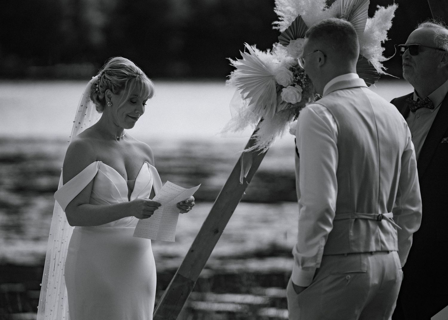 Bride reads vows to groom at outdoor waterfront wedding ceremony in black and white photo.