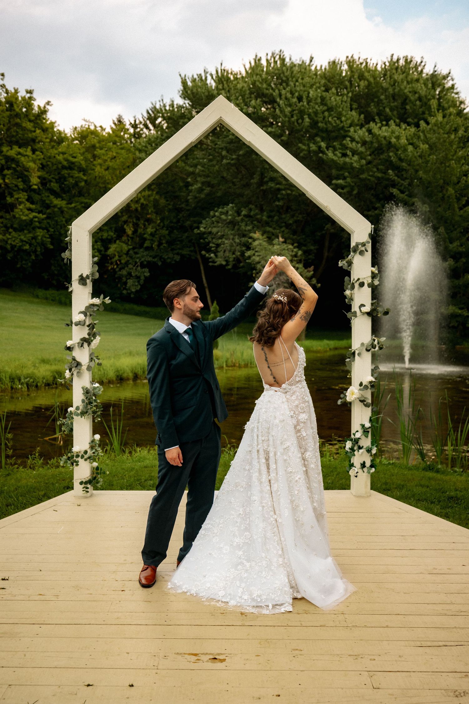 Bride and groom dancing under a white arch on a wooden deck with a fountain and lush greenery in the background.