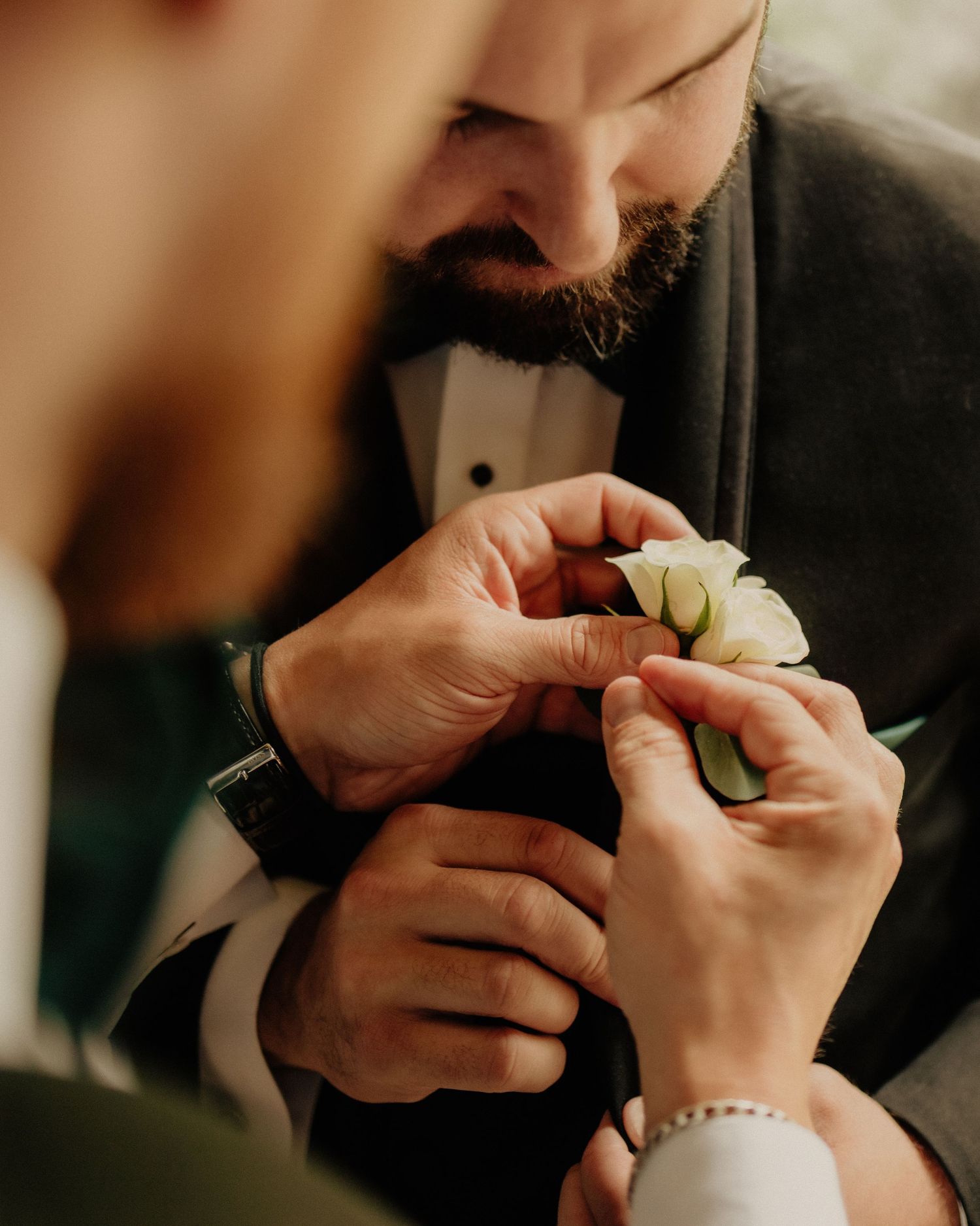 Groom in tuxedo having white rose boutonniere pinned to lapel at wedding.